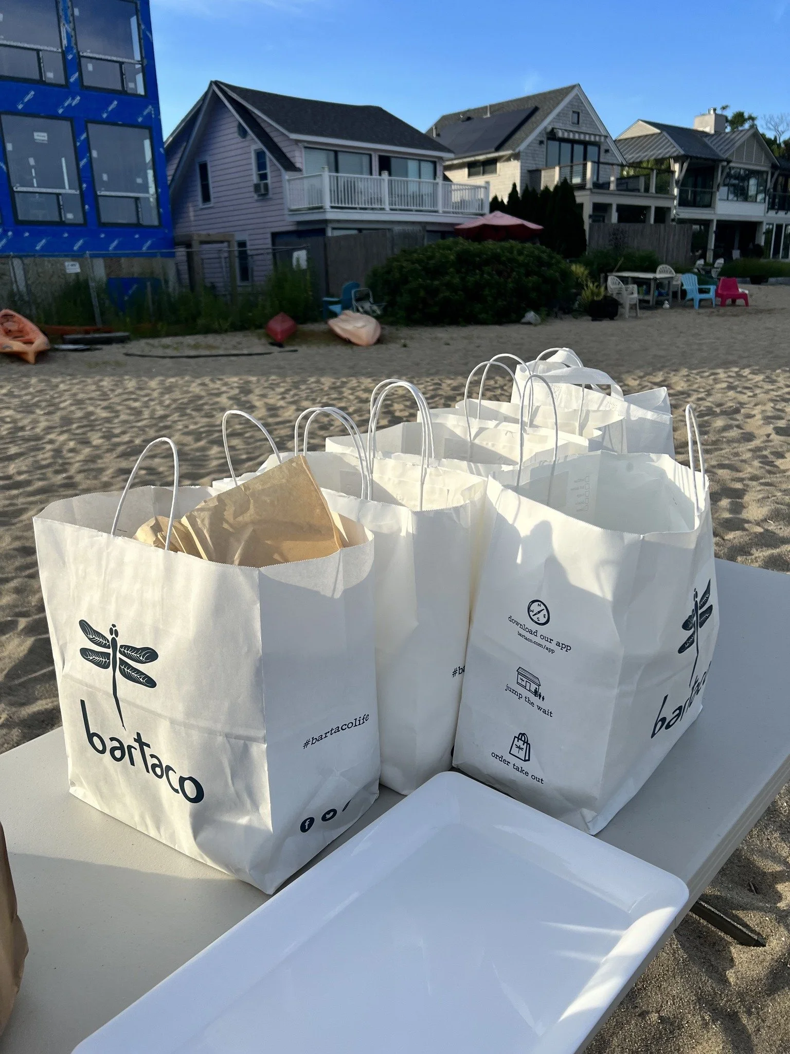 Three white paper bags with restaurant branding, sitting on a table on a sandy beach with picnic tables, houses, and kayaks in the background.