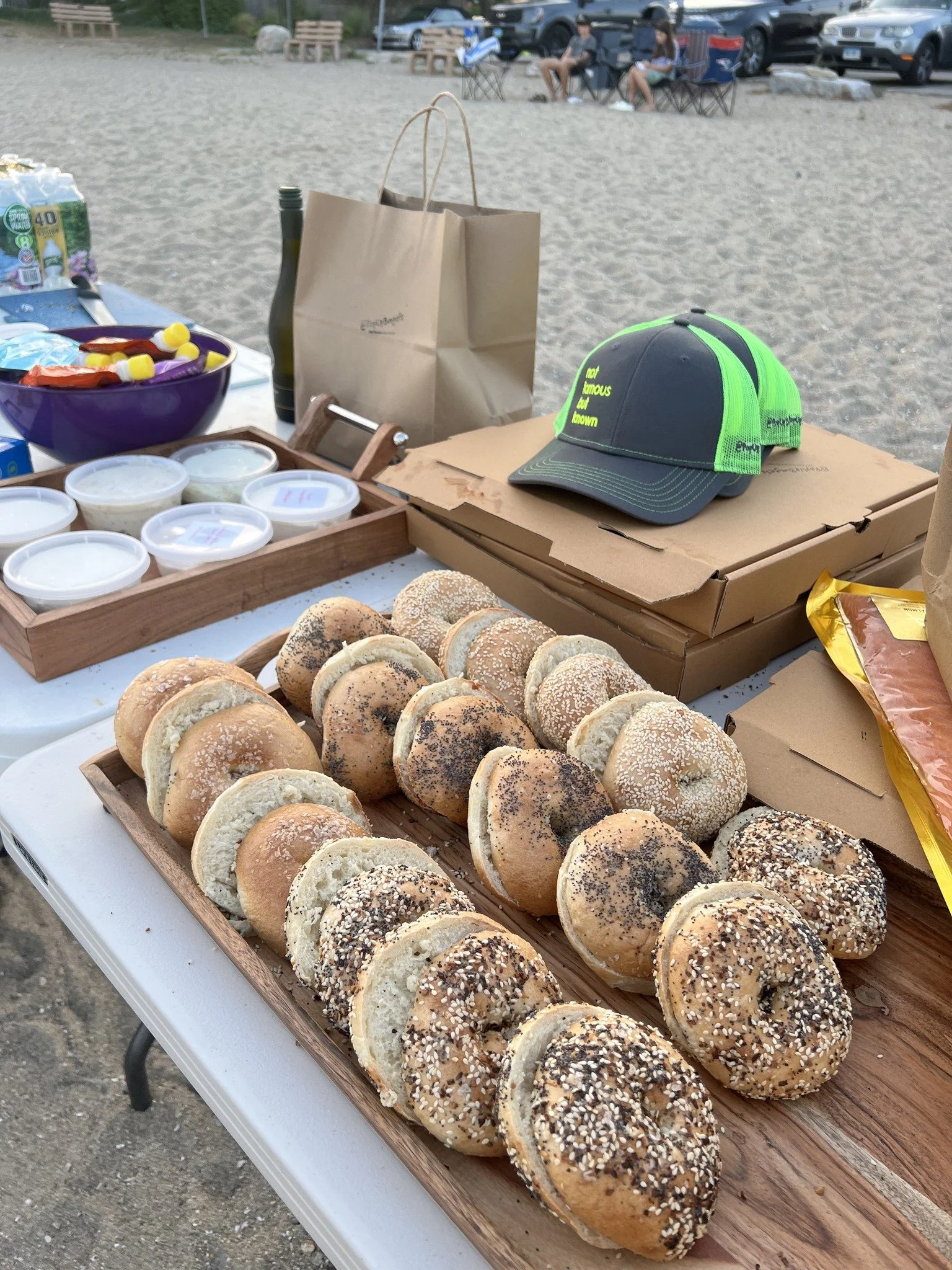 Books on the Beach 203 by Carrie Kerner A table of assorted bagels with toppings such as sesame and poppy seeds, at an outdoor gathering on a sandy area with parked cars in the background. PopUp Bagels
