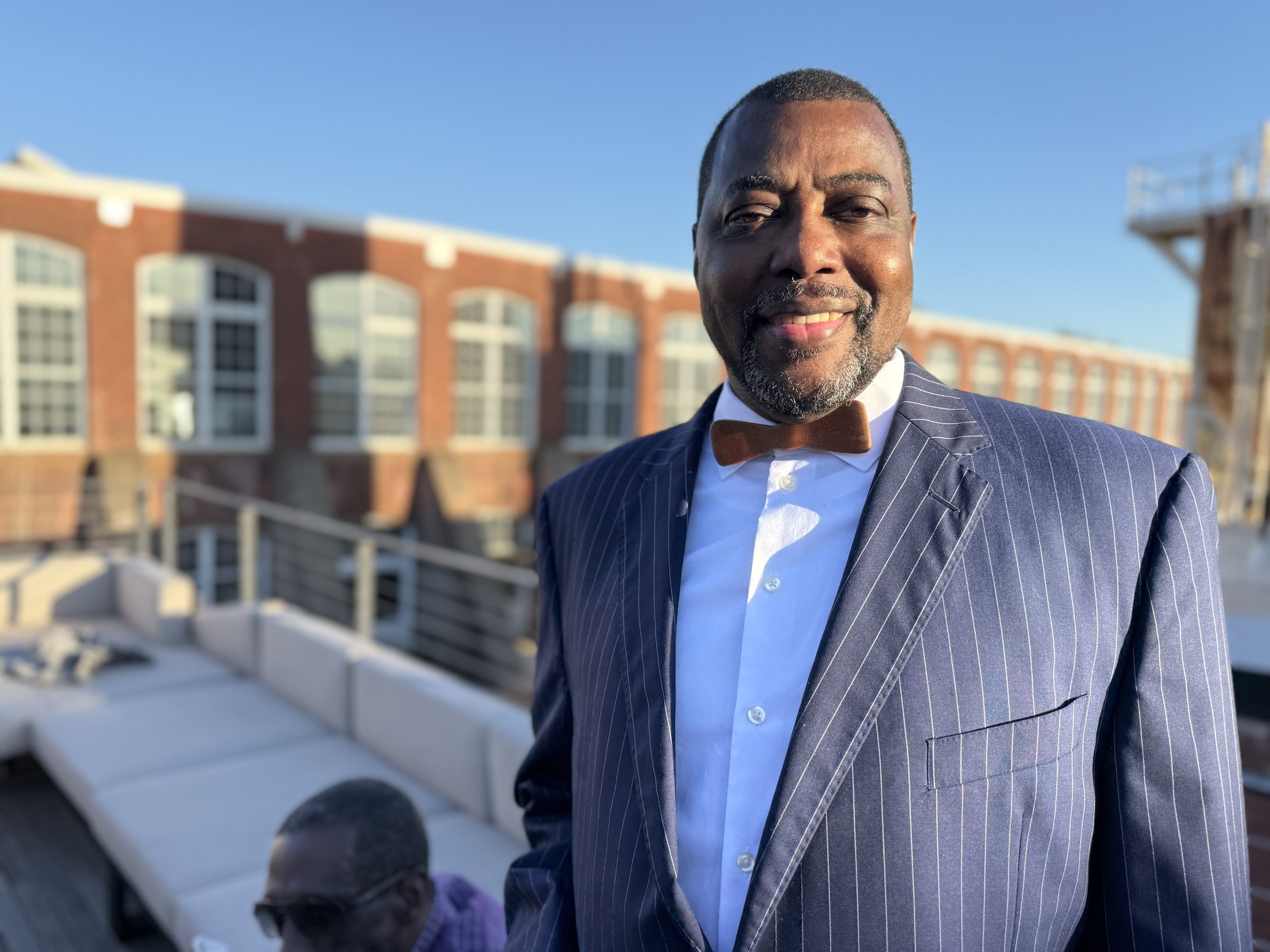 A man in a navy pinstripe suit with a white shirt and brown bow tie, smiling outdoors on a rooftop with a brick building and blue sky in the background.