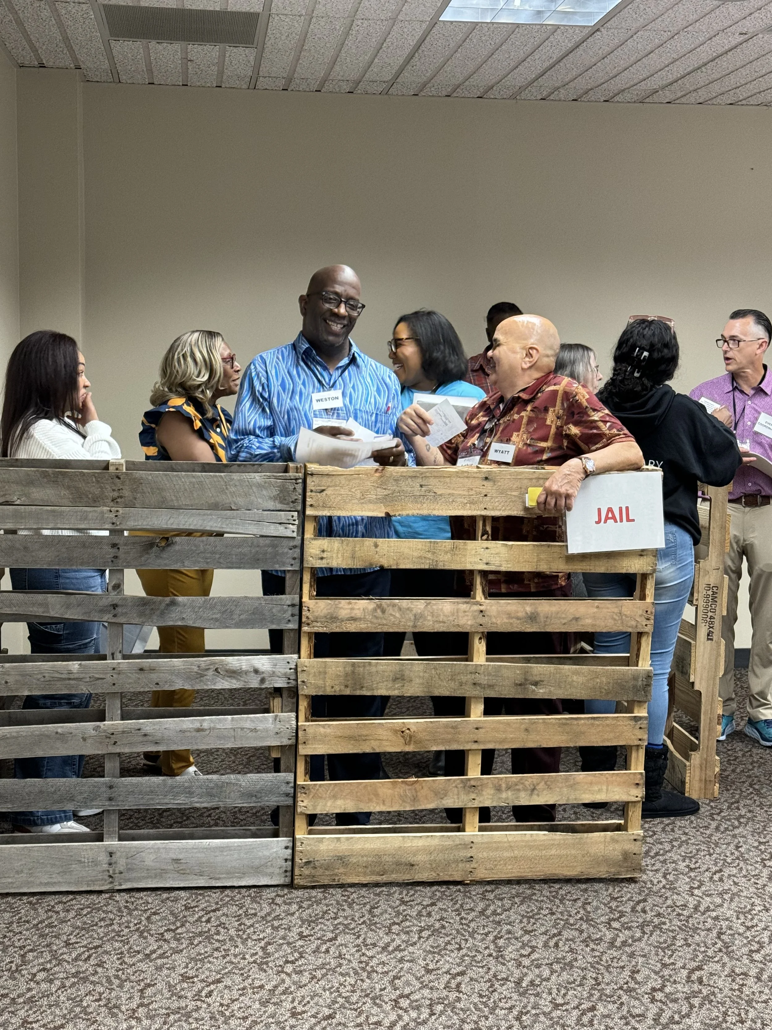 Group of diverse adults socializing and talking at an event, with some standing behind a wooden barrier that has a 'JAIL' sign on it.