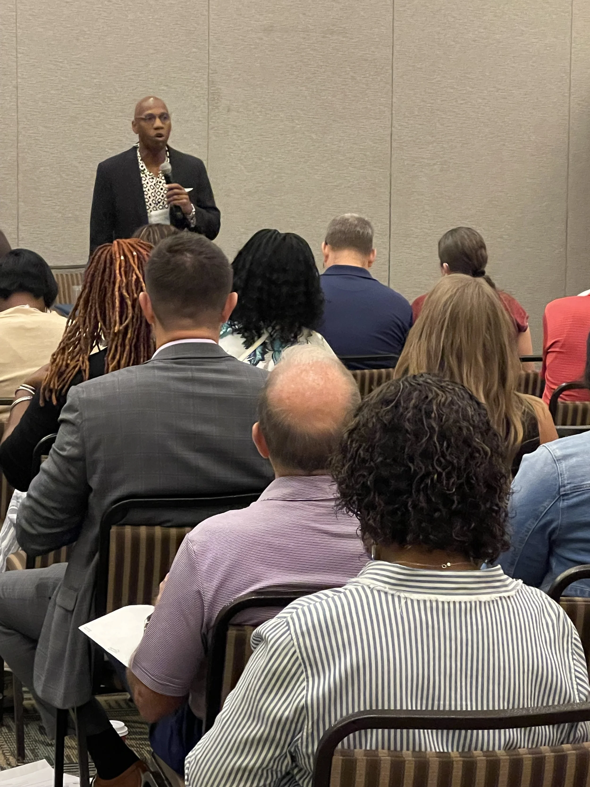 A man wearing a dark suit speaking into a microphone in a conference room with an audience facing forward.