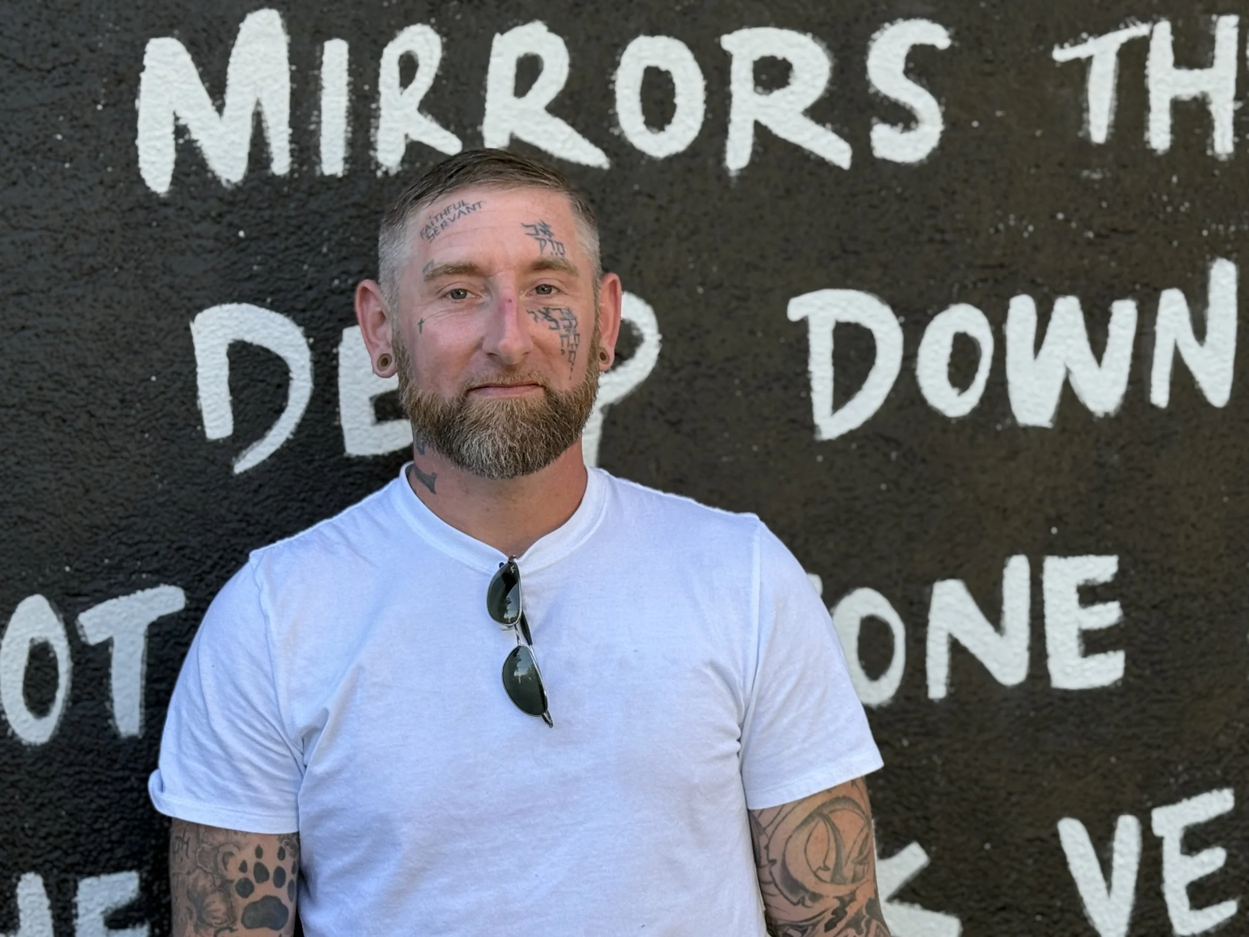 A man with tattoos and a beard standing in front of a black wall with white painted words.