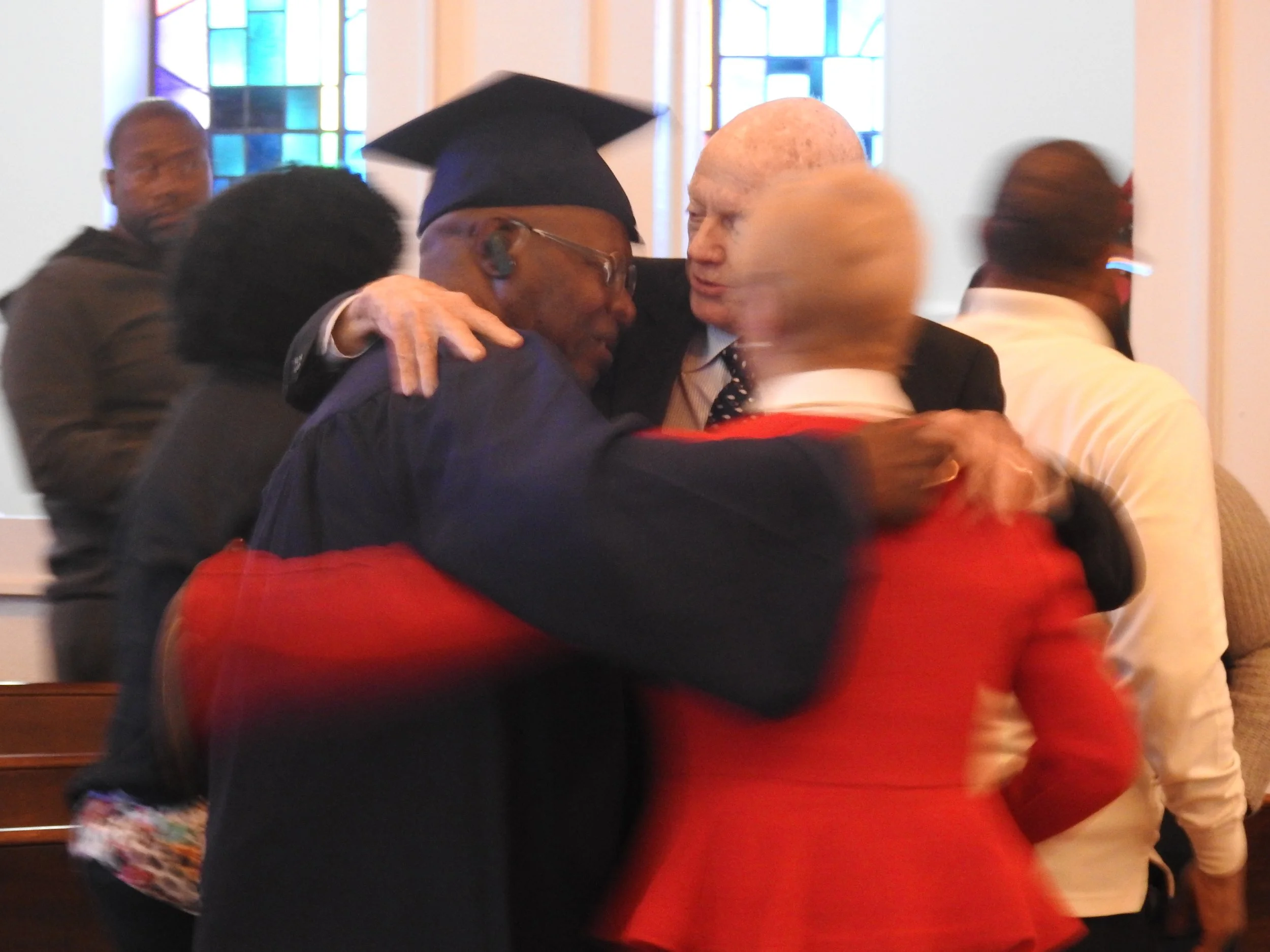 Mentor and his wife hugging a Soteria graduate in what appears to be a church or chapel, with stained glass windows in the background.