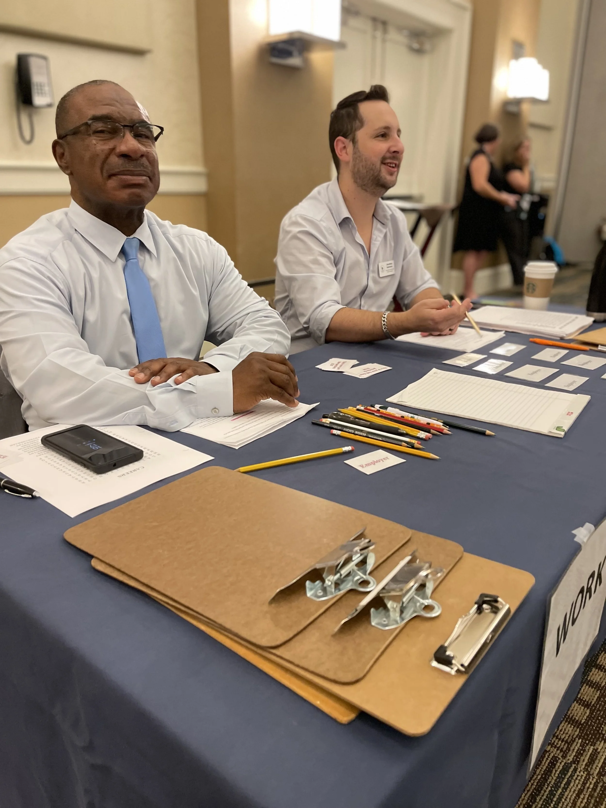 Two men sit at a table with papers, pens, and clipboards, smiling at a meeting or conference in a room with beige walls and lights.