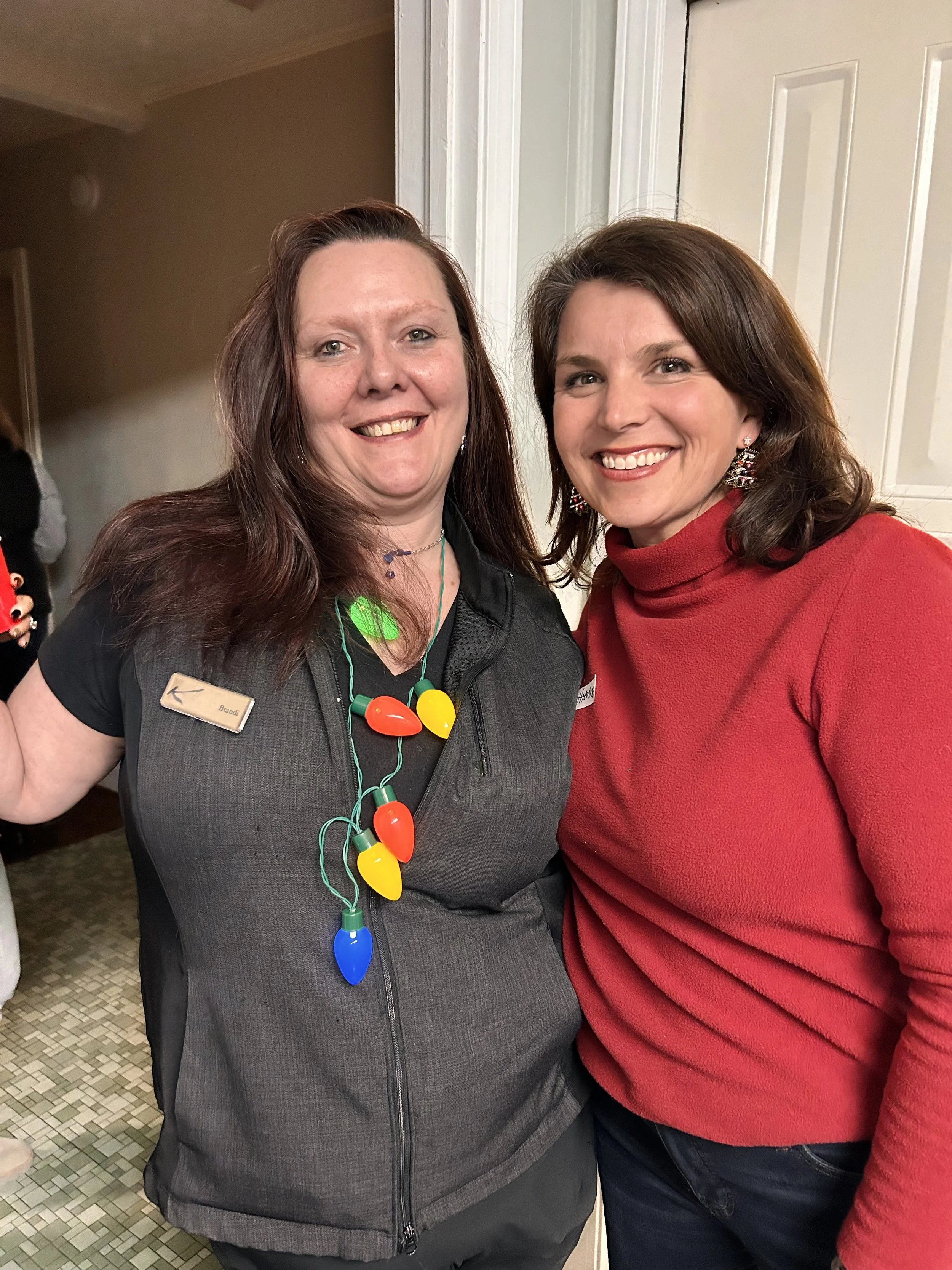 Mentor and Mentee smiling, one wearing a holiday necklace, the other in a red turtleneck sweater, standing in an indoor setting.