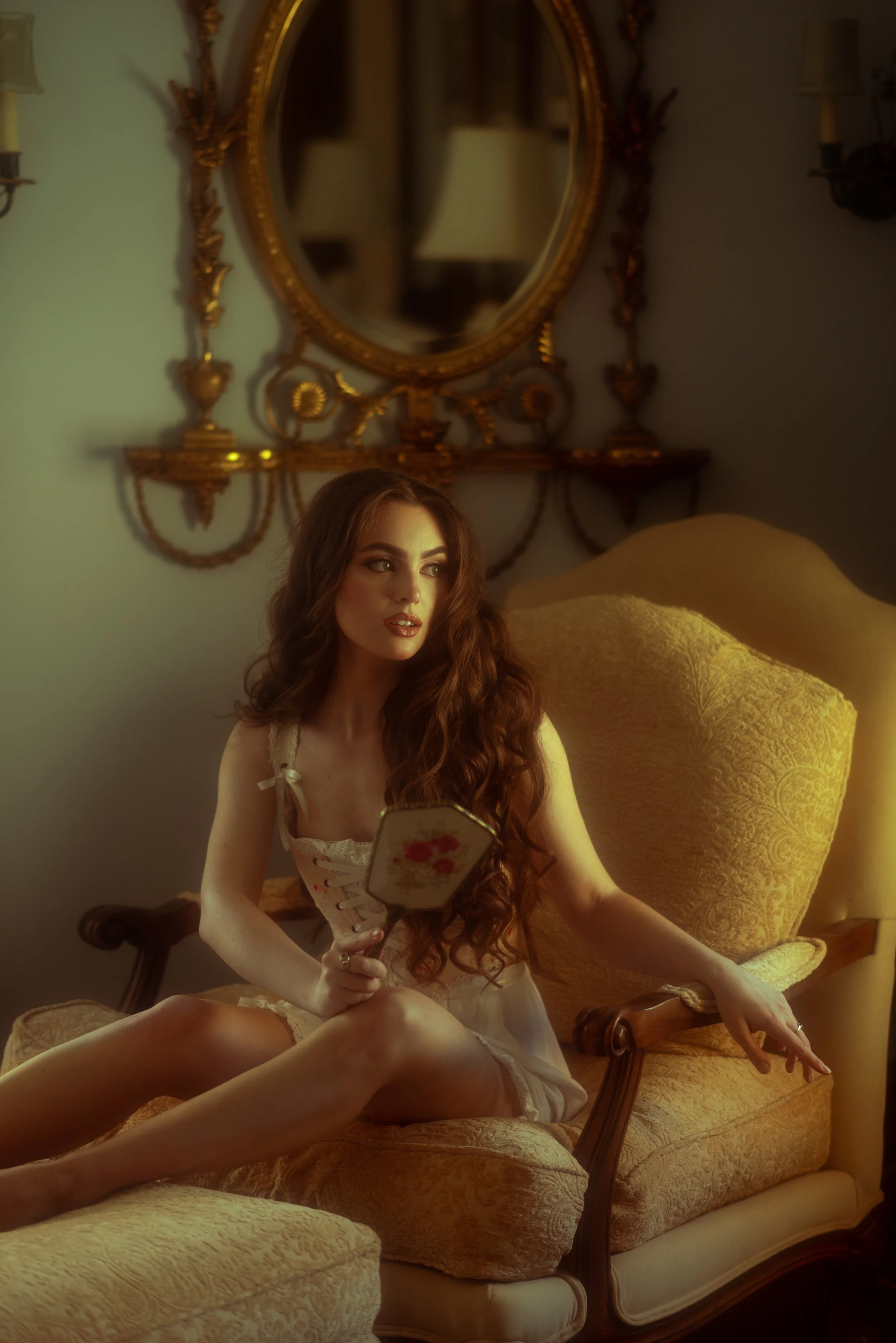 A woman with long, curly red hair sitting on an ornate yellow velvet armchair, holding a hand mirror, in a dimly lit vintage-style room with a large gilded mirror and wall sconces.