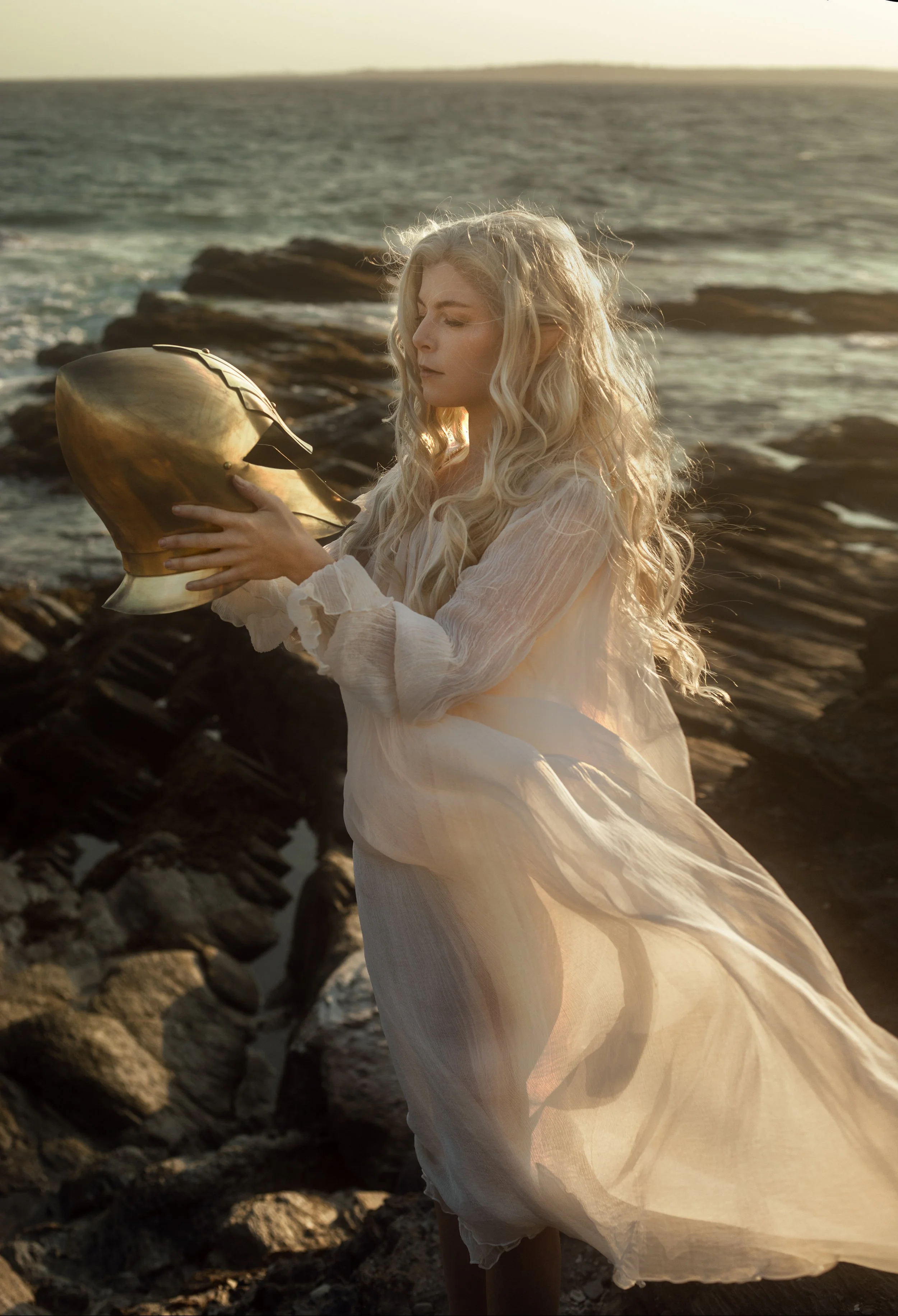 A woman with long, curly blonde hair wearing a flowing, sheer white dress stands on a rocky shoreline holding a metallic helmet against a sunset background.