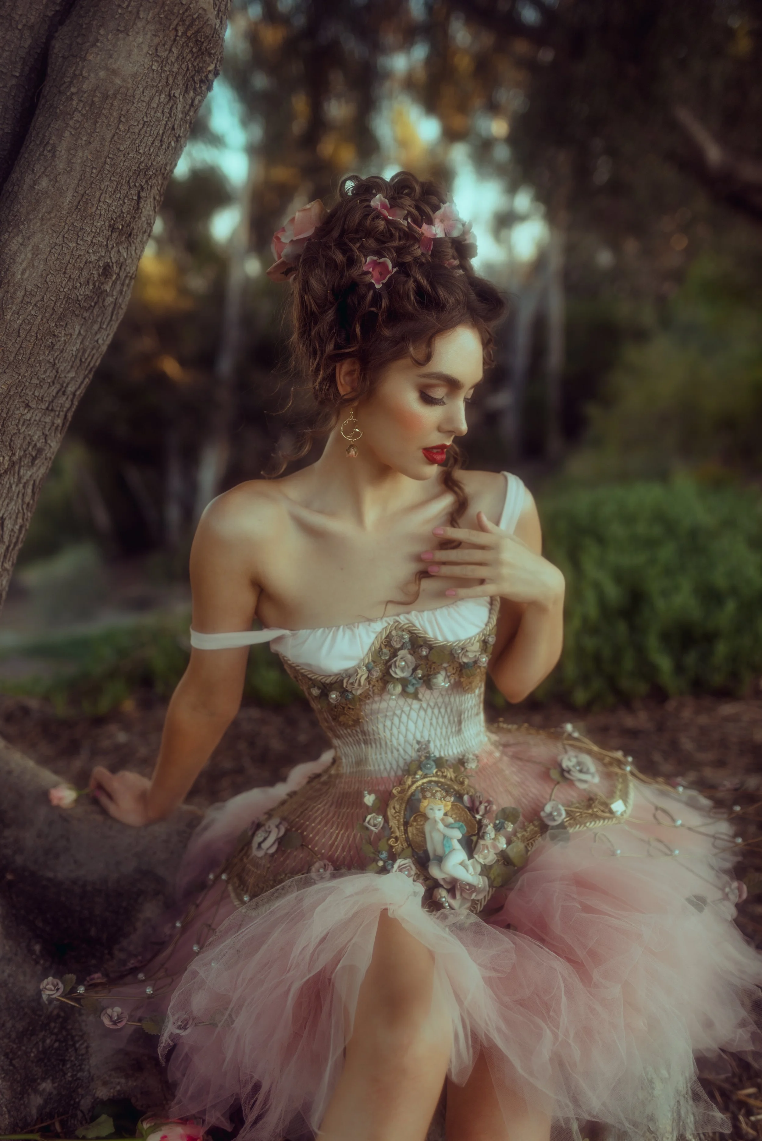 A woman with elaborate curly hair adorned with pink flowers, wearing a decorative corset and a pink tulle skirt, sitting outdoors near a tree in a wooded area, with a contemplative expression.