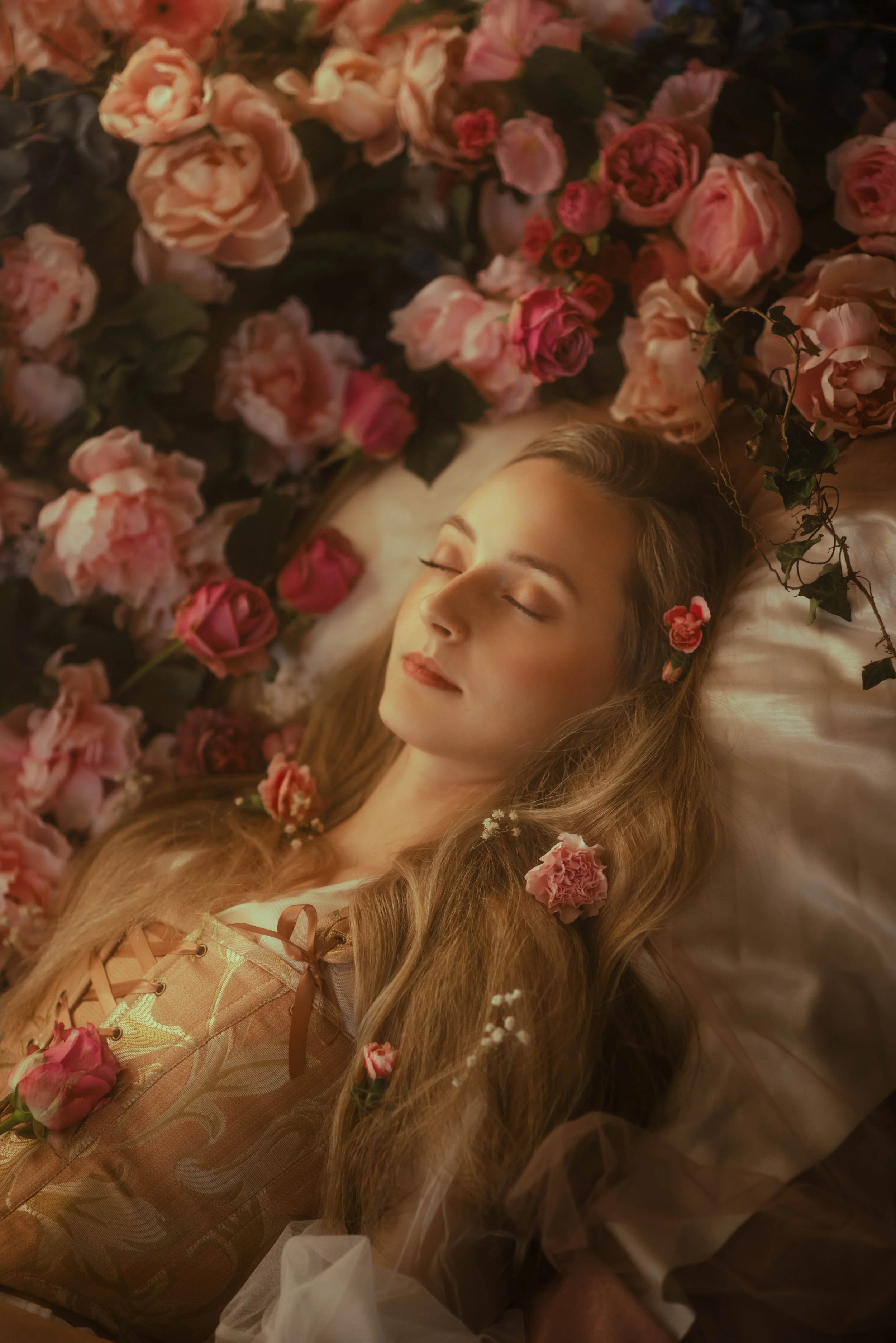 A woman with long, wavy hair and light makeup lies on a bed surrounded by pink and red roses. She has her eyes closed and appears peaceful, with a small flower in her hair and flowers in her hair and on her dress.