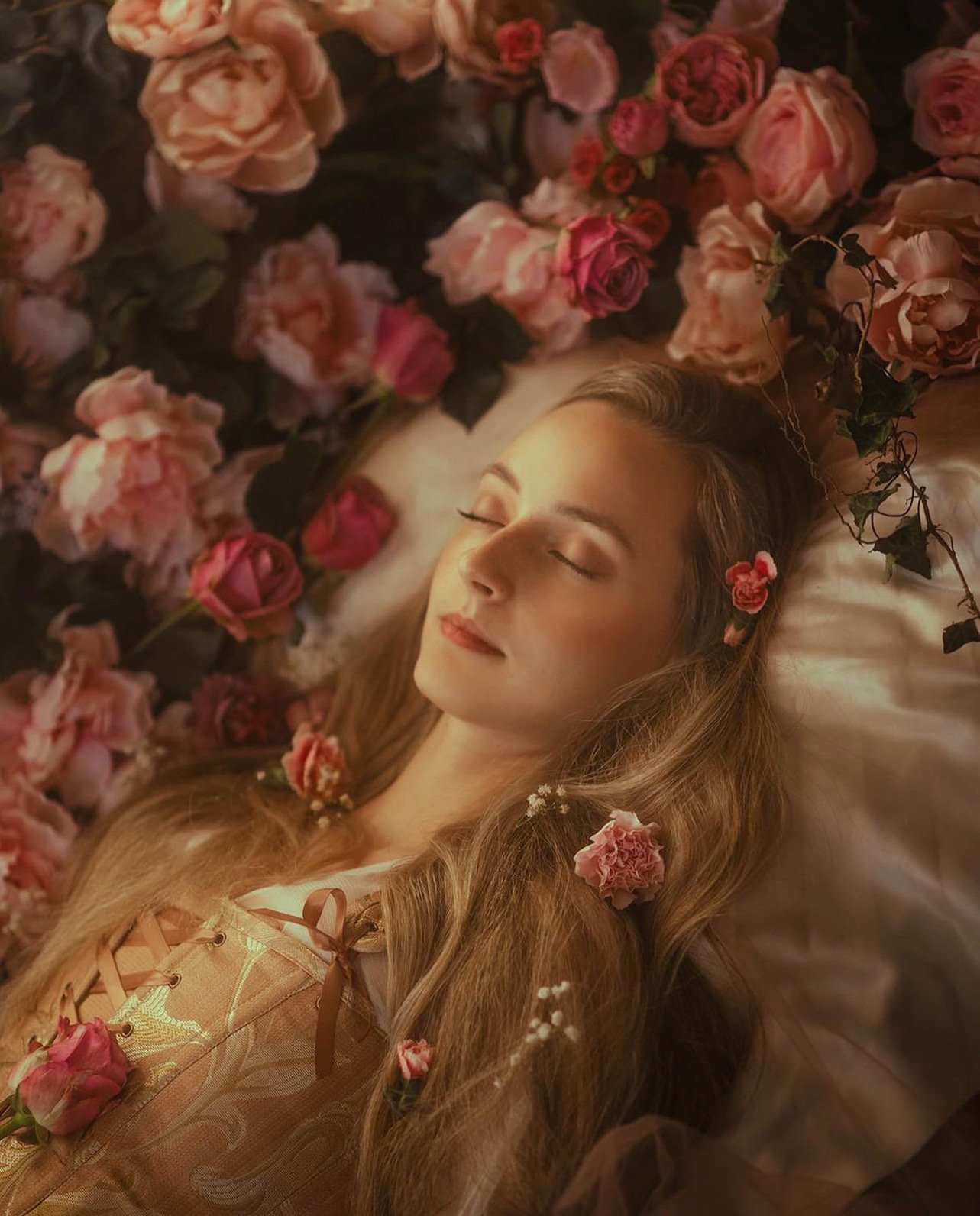 A woman with long blonde hair is peacefully sleeping on a bed surrounded by pink and peach roses, with some flowers resting on her hair and body.