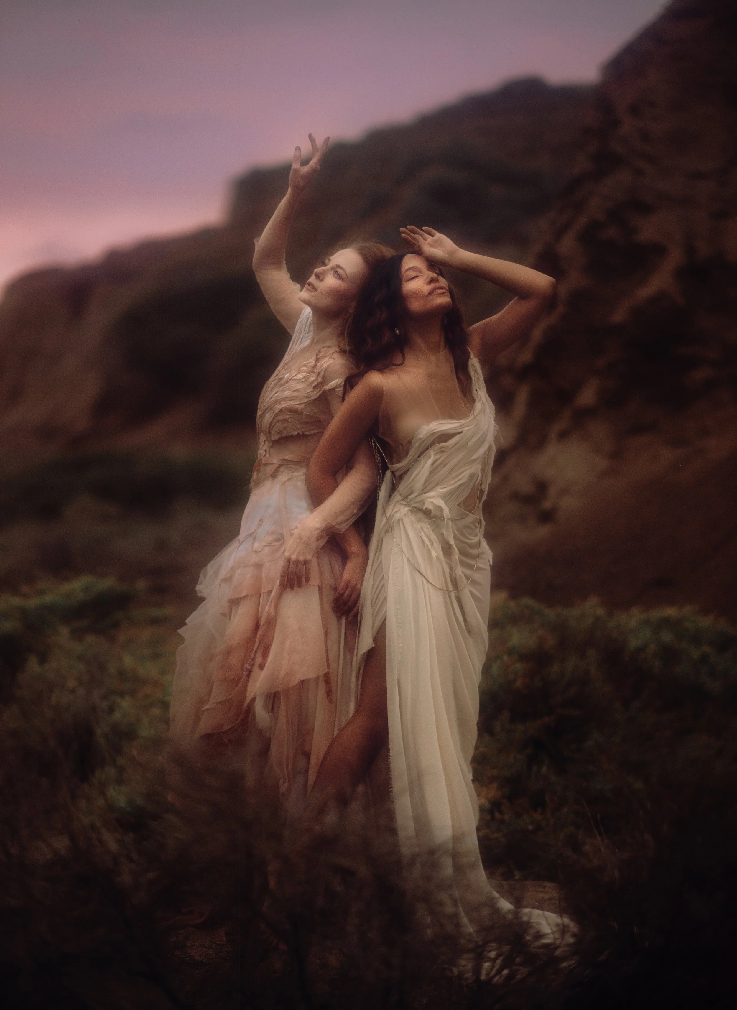 Two women in flowing dresses posing outdoors at sunset with a rocky hillside in the background.