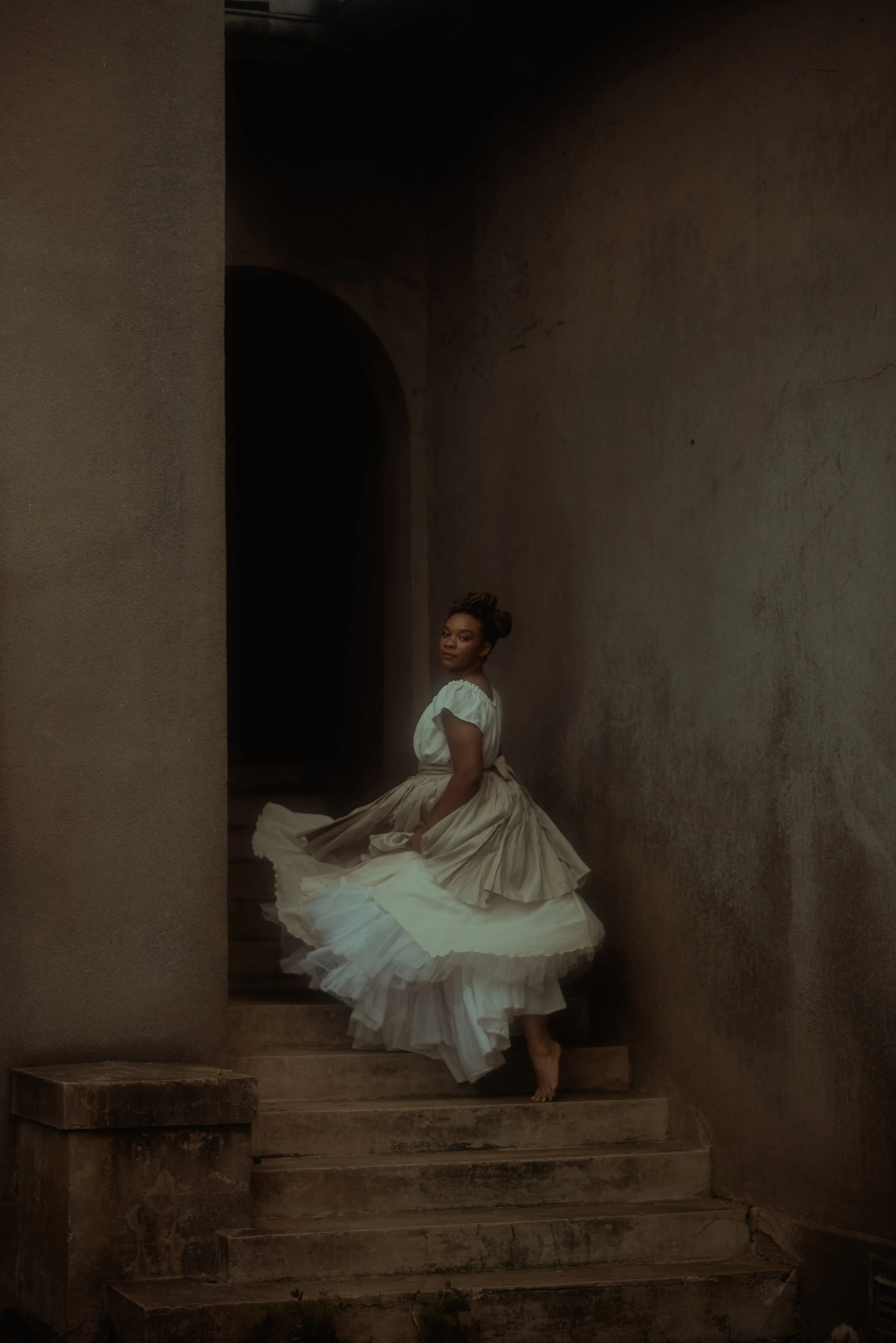 A woman in a vintage dress standing on stone steps in a dark, narrow passageway.