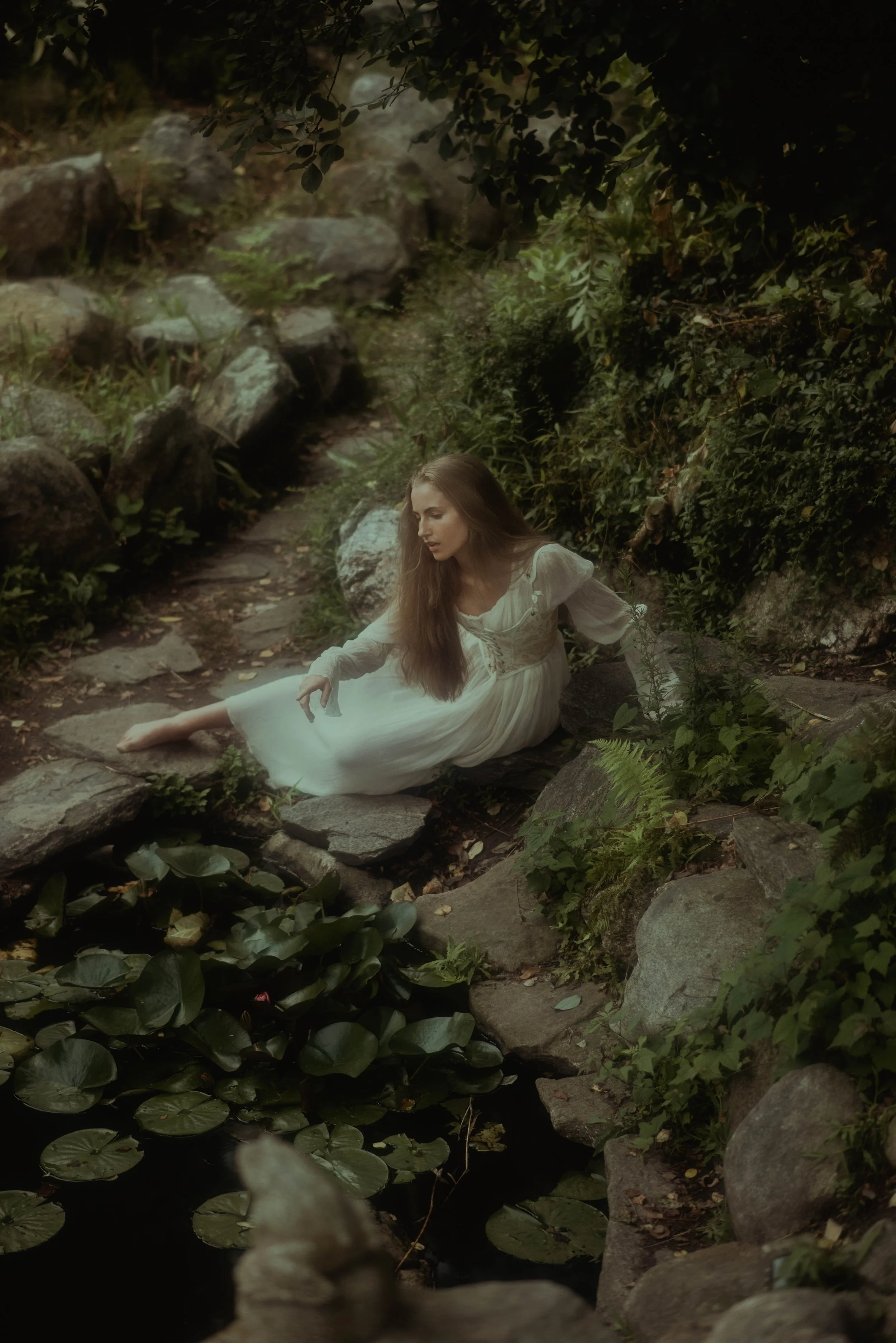 A woman in a white dress sitting on a rock in a lush, green forest by a pond with lily pads.