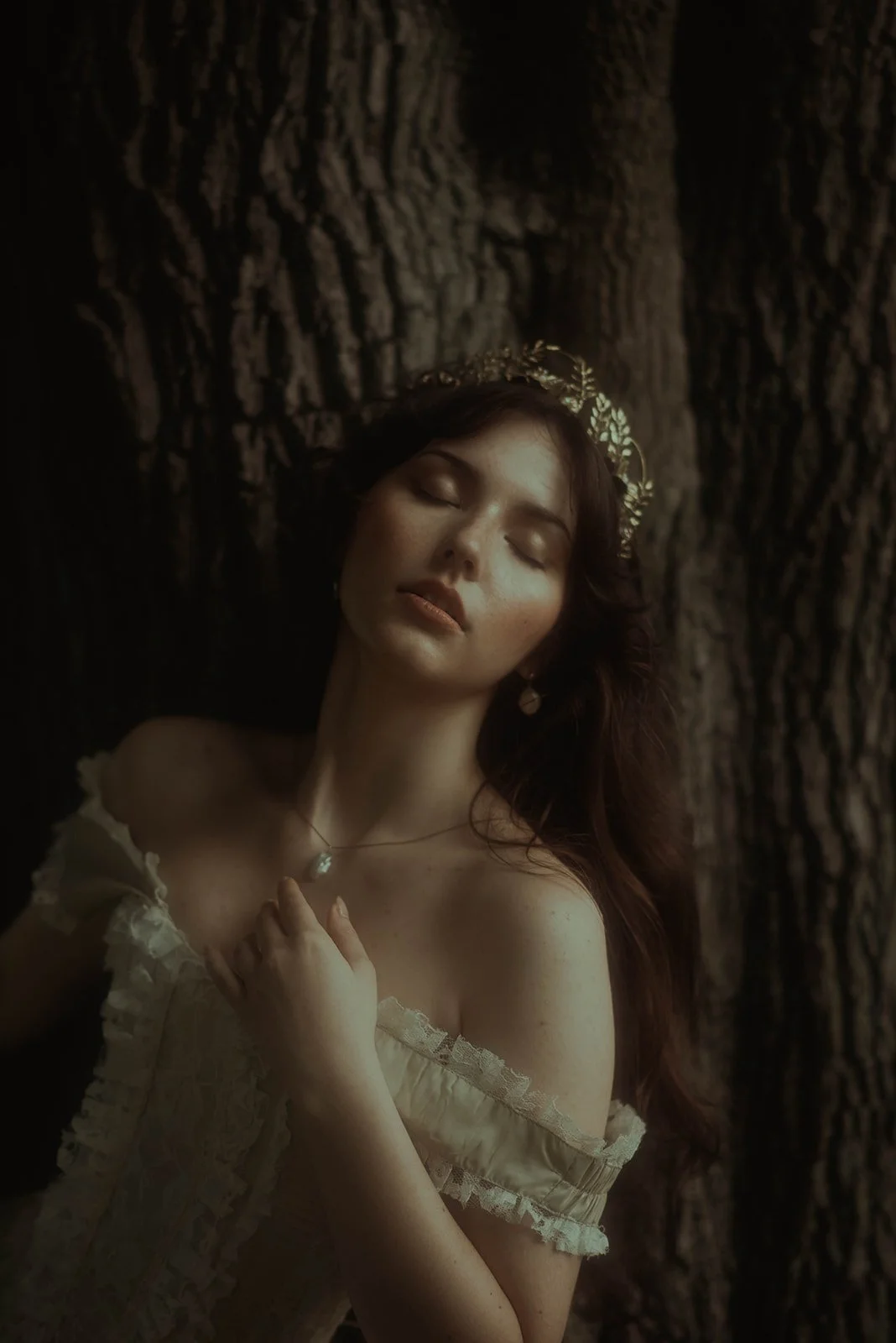 A woman with closed eyes and long brown hair, wearing a floral headband and a cream off-shoulder dress, stands next to a large tree trunk in a dark setting.