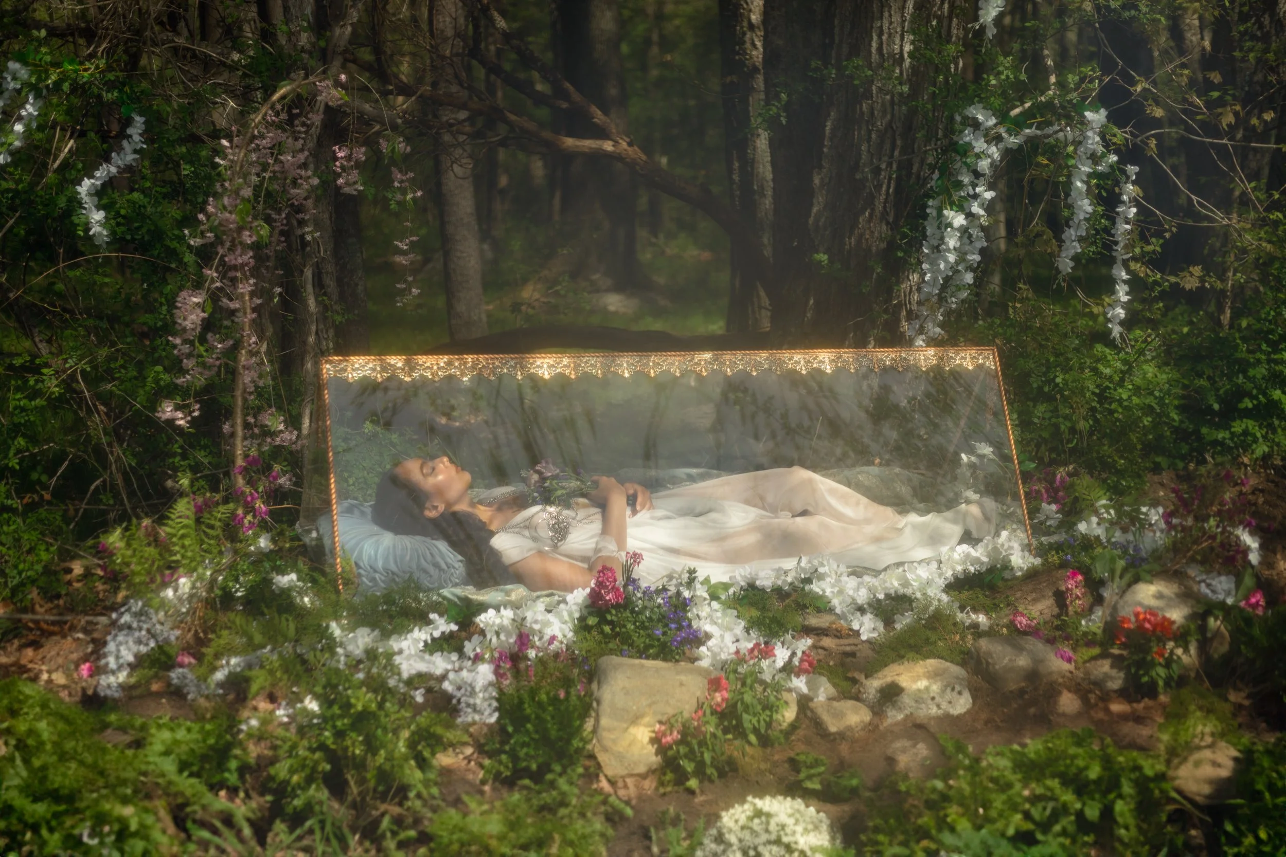 A woman lying on a bed surrounded by flowers and greenery in a forest, with a transparent canopy over her.