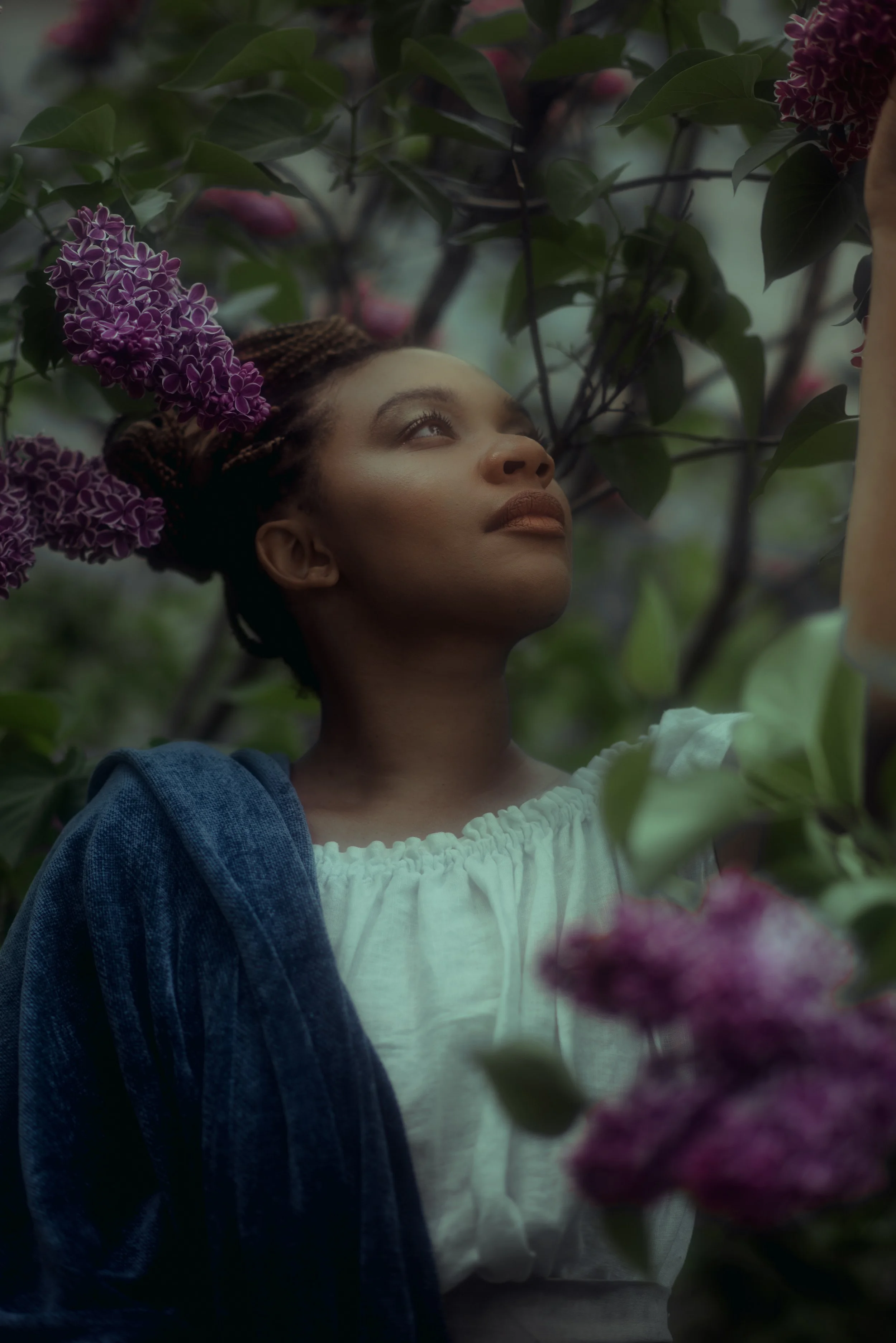 A woman with braided hair looking up among pink and purple flowers and green leaves.