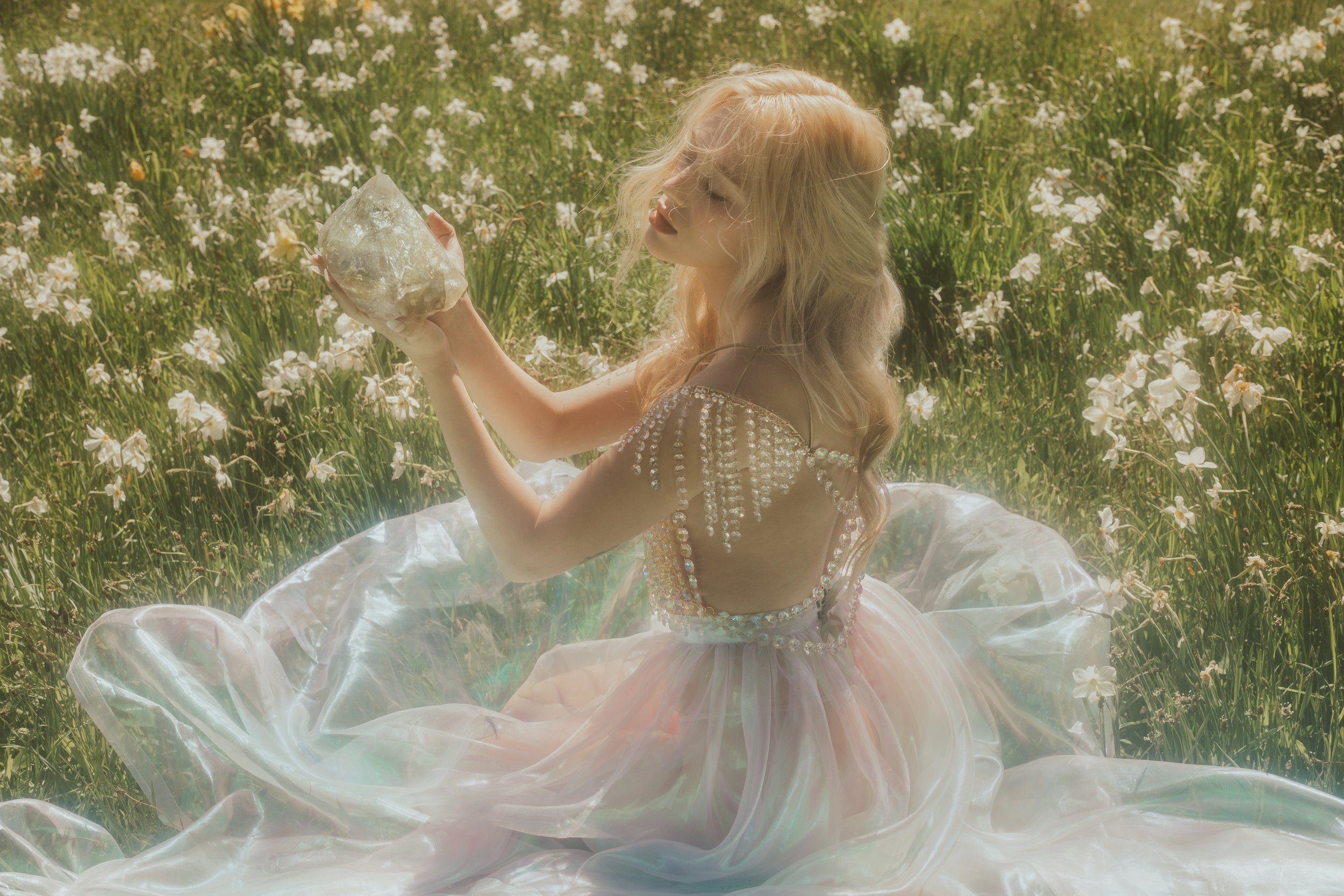 A woman with long blonde hair in a pearl-studded dress sitting in a field of white flowers, holding a large crystal.