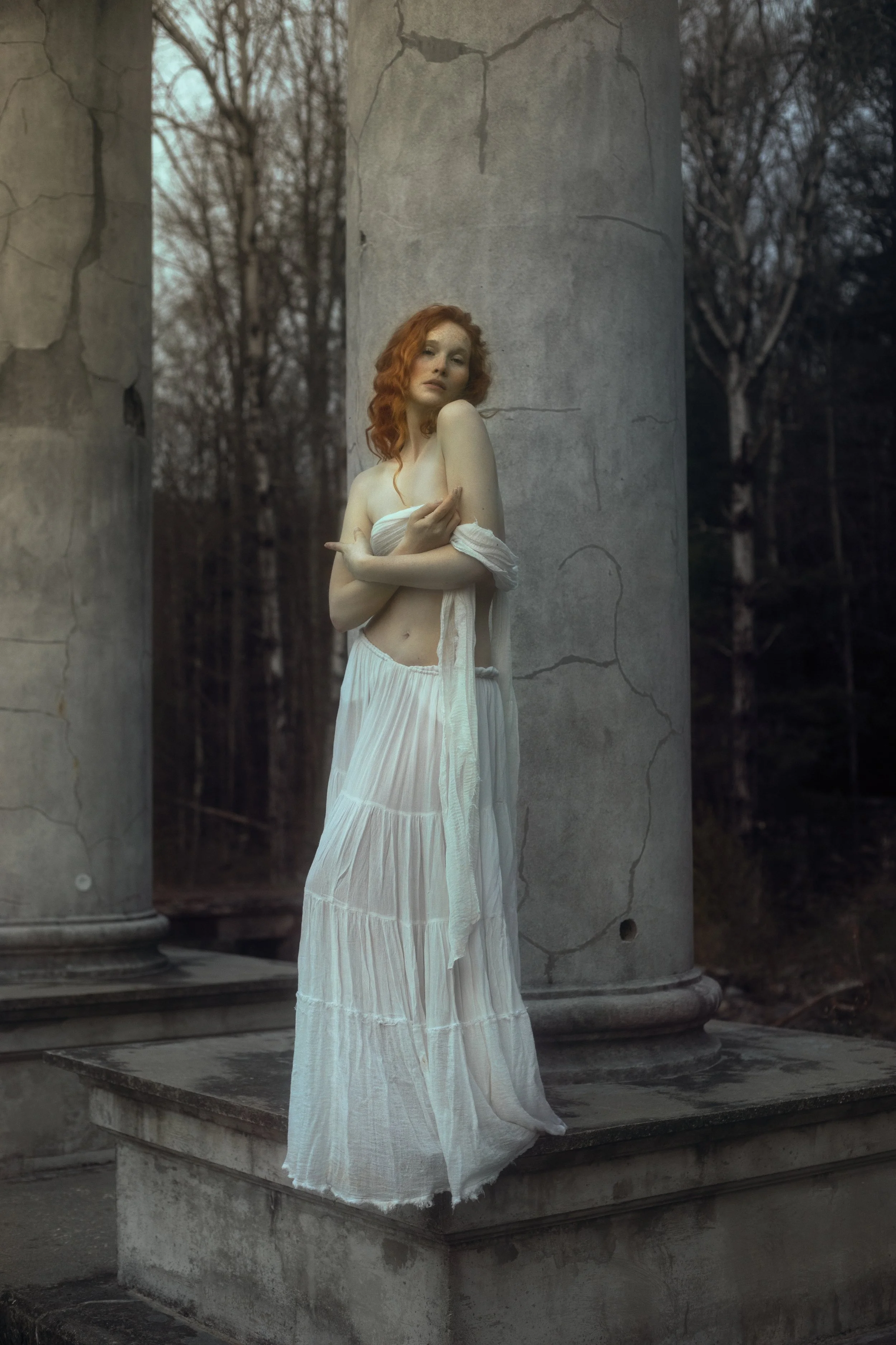 A woman with red hair wearing a white skirt and wrap is standing between two broken concrete pillars outdoors, with a forest in the background.