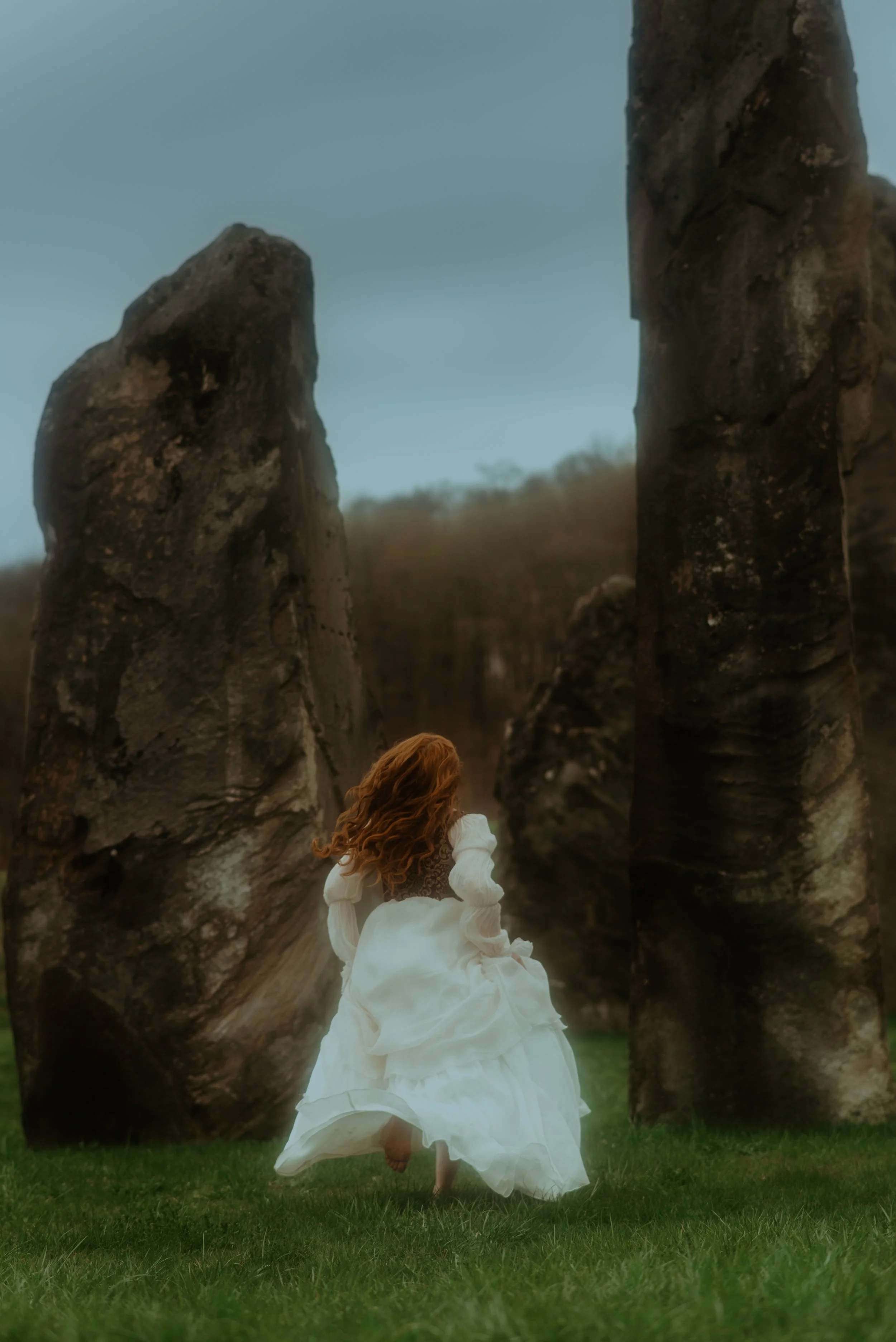 A woman in a long white dress with reddish hair kneeling on green grass between two large standing rocks or stone monoliths.