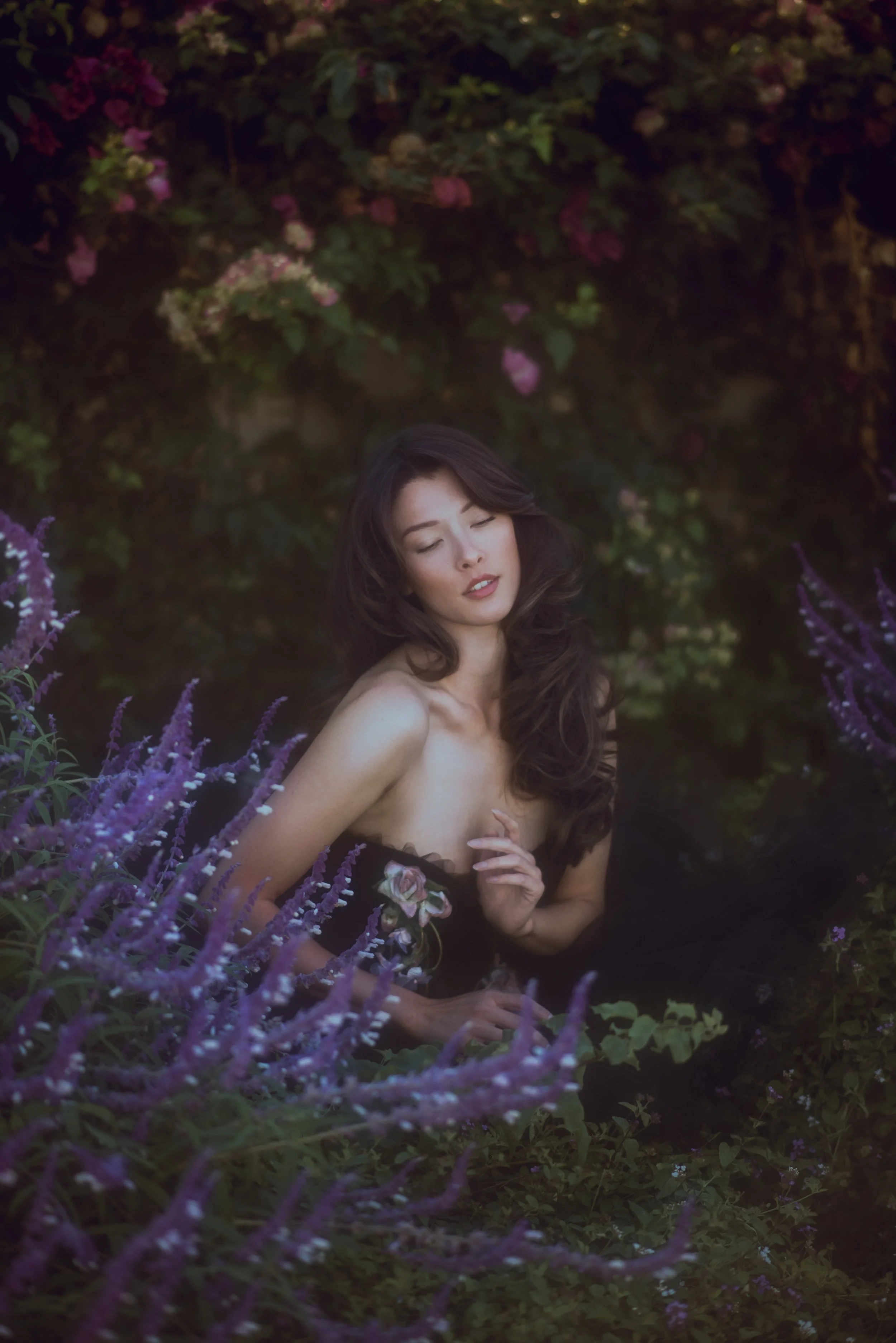 A woman with long dark hair surrounded by various purple and pink flowers, sitting in a garden or natural setting with her eyes closed and a peaceful expression.