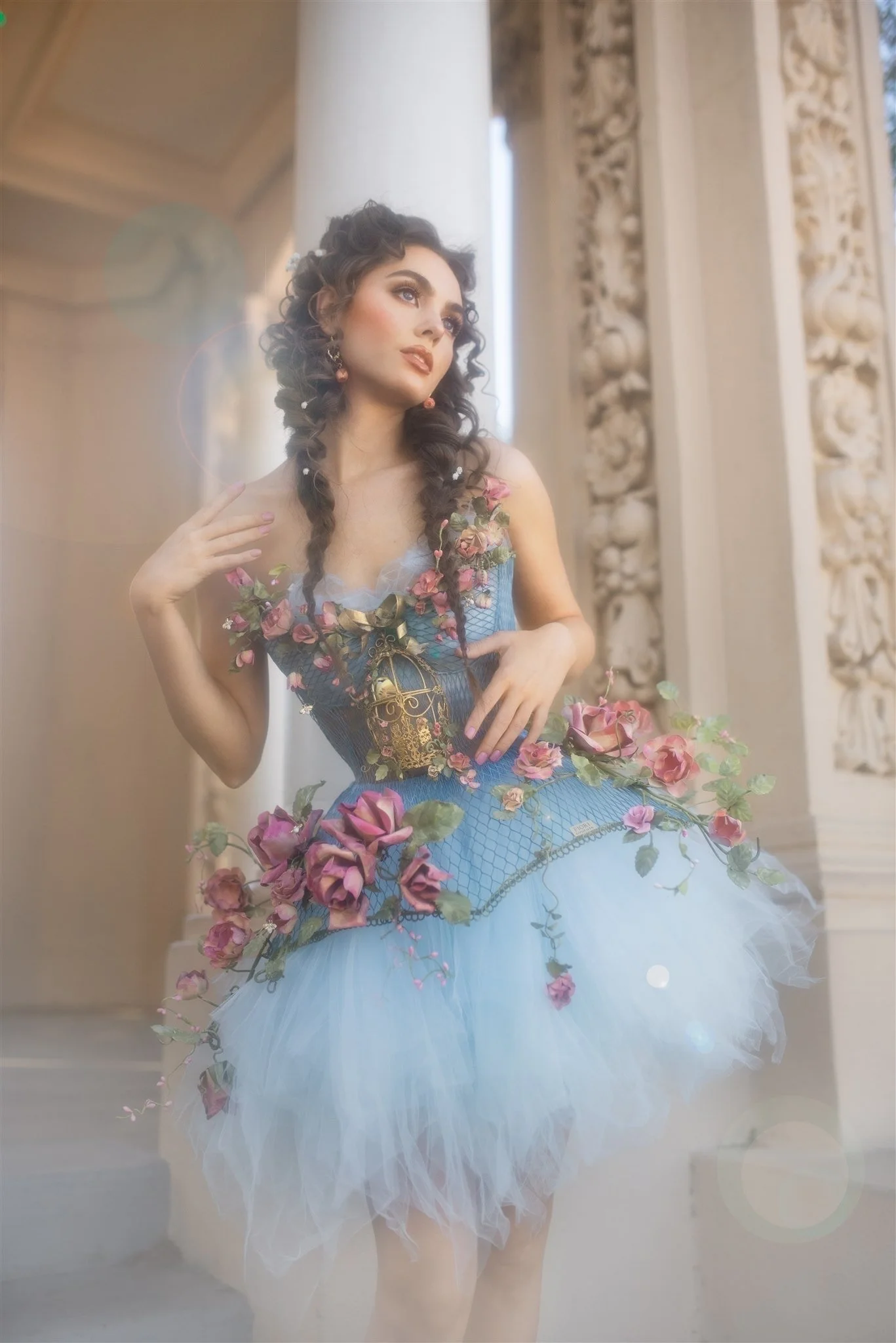 Woman with curly hair in a blue dress adorned with pink roses and a cage design, posing in front of ornate architecture.
