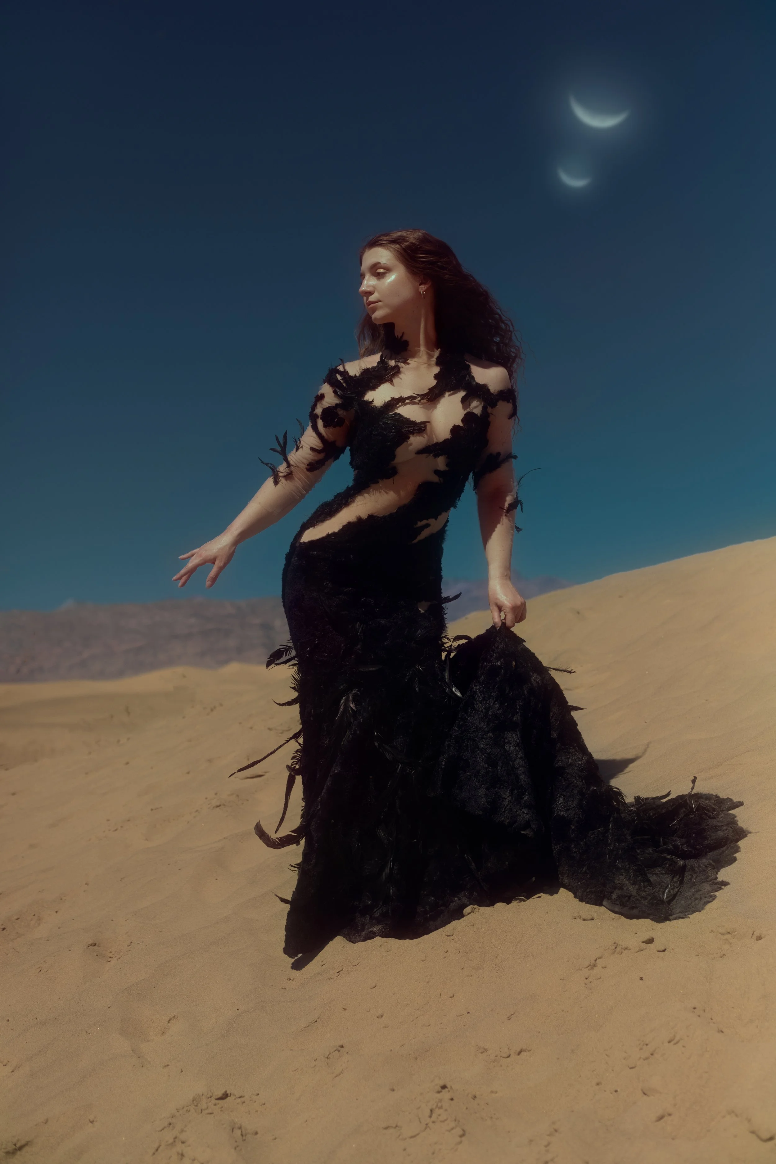 A woman in a black dress standing in a desert landscape under a dark blue sky with a crescent moon.