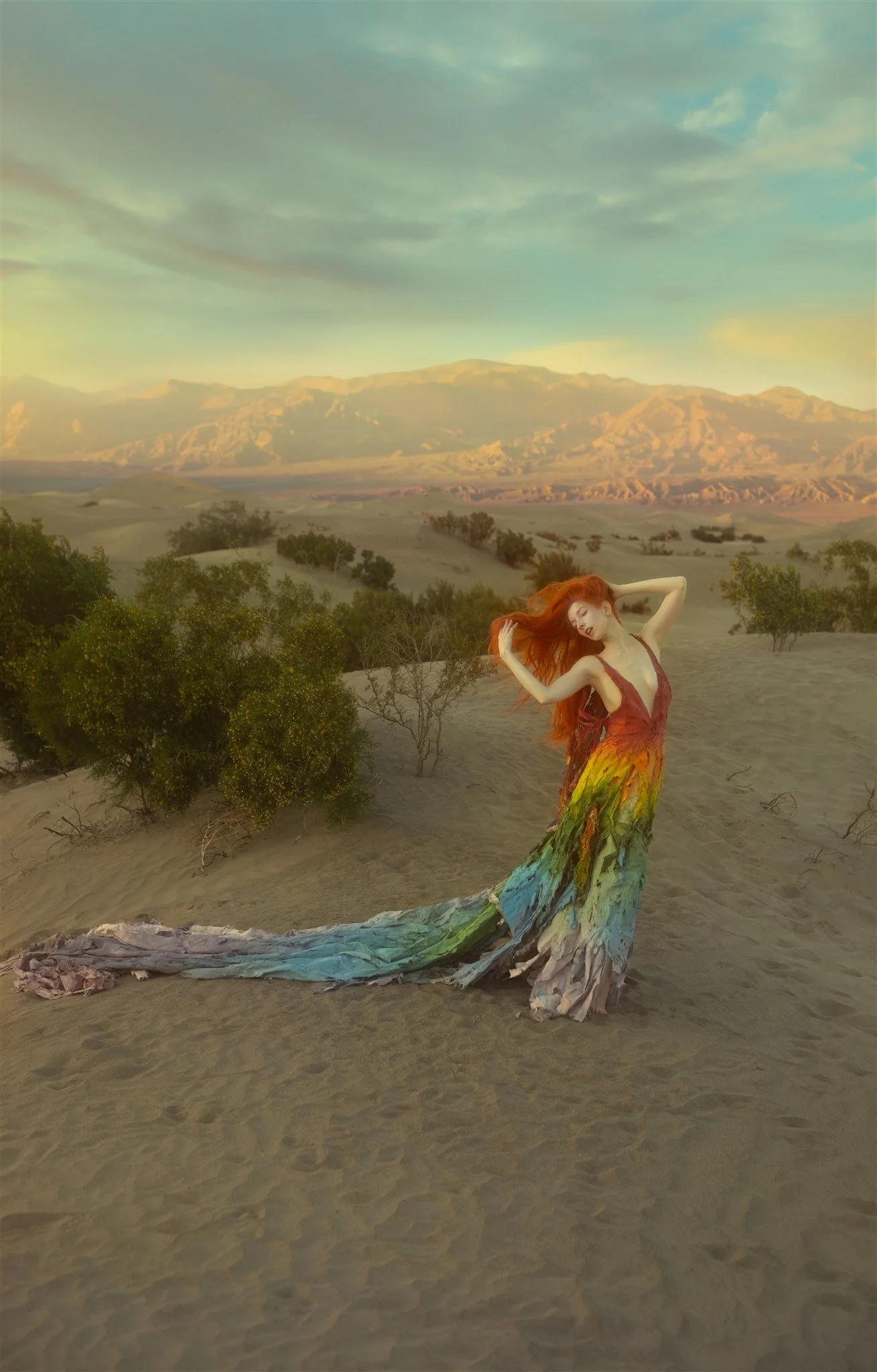 A woman with long red hair in a colorful dress poses in a desert landscape with mountains in the background and the sky partly cloudy.