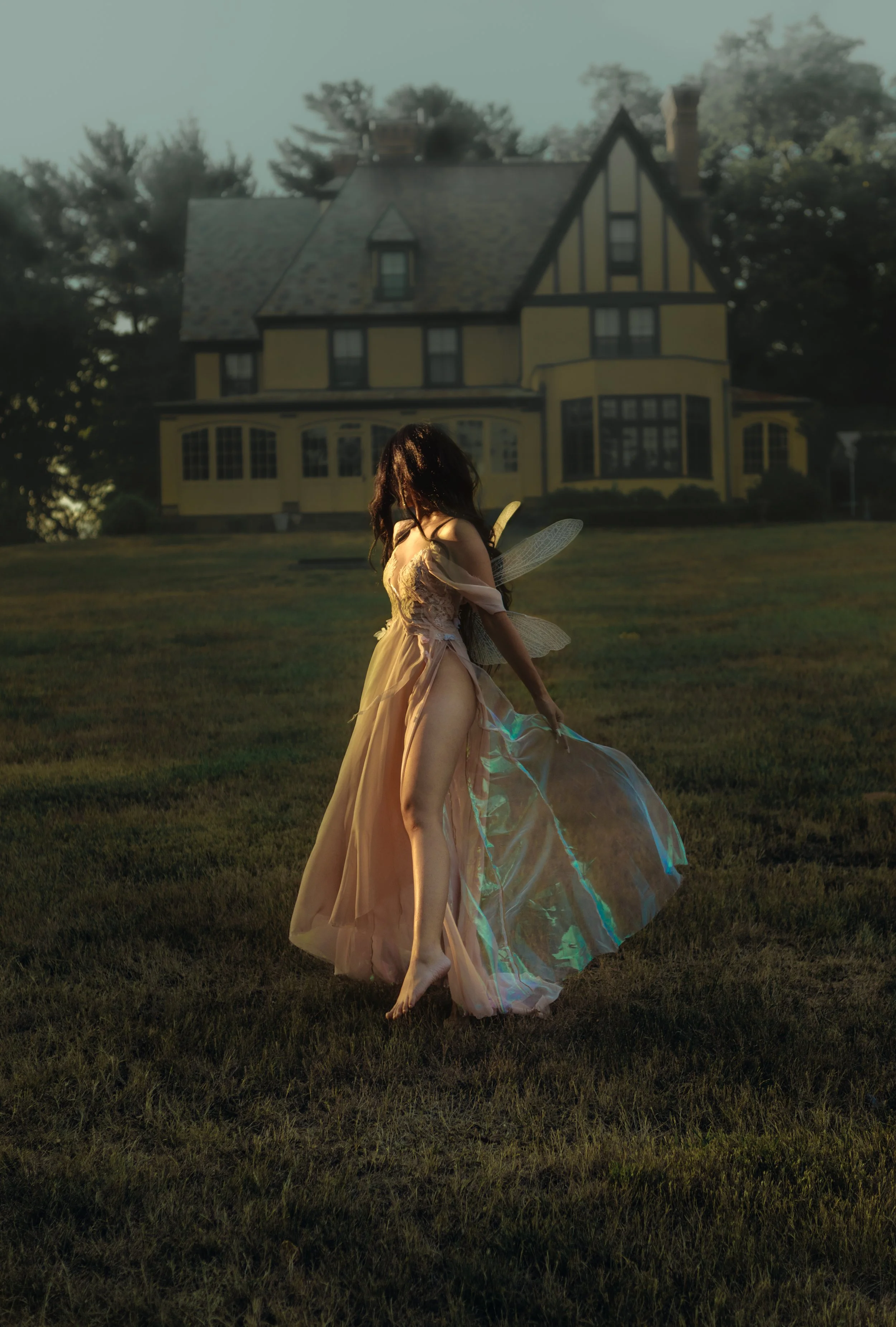 A woman with fairy wings wearing a flowing dress, holding a fan in front of a large house at sunset.