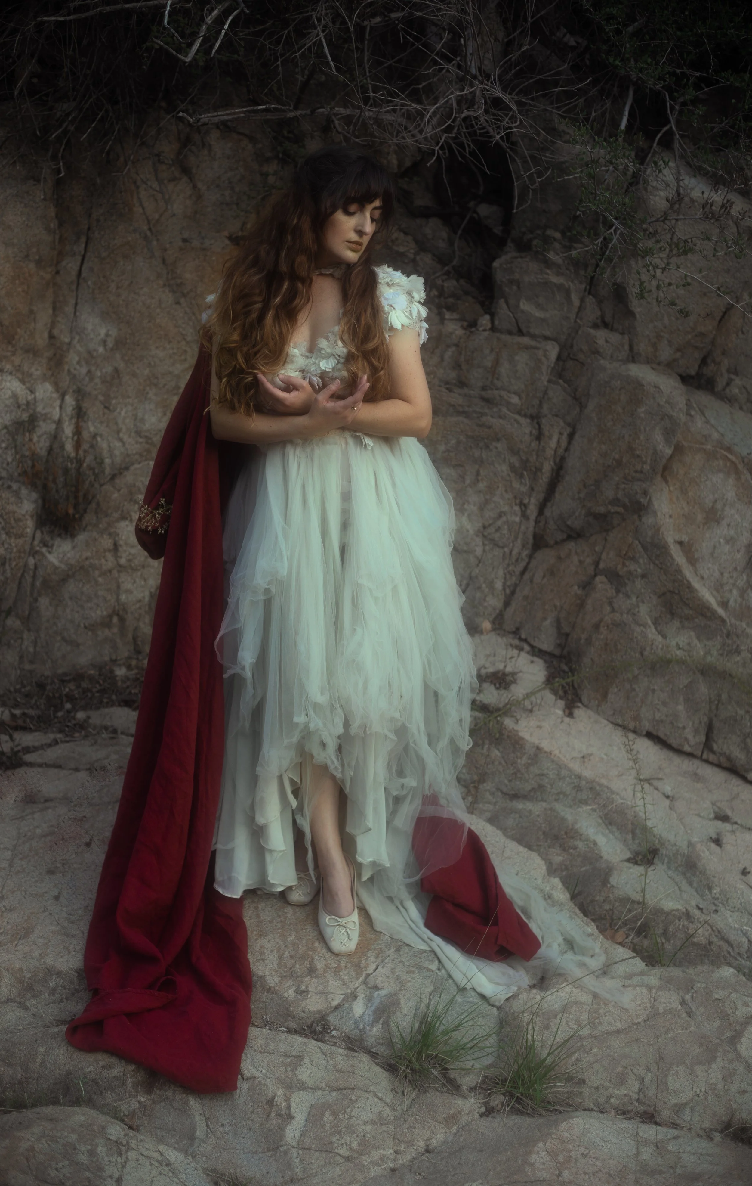 A woman dressed in a vintage white gown with tulle and floral details, standing barefoot on a rocky terrain, with a red cloak draped over her shoulder, gazing downwards with a contemplative expression.