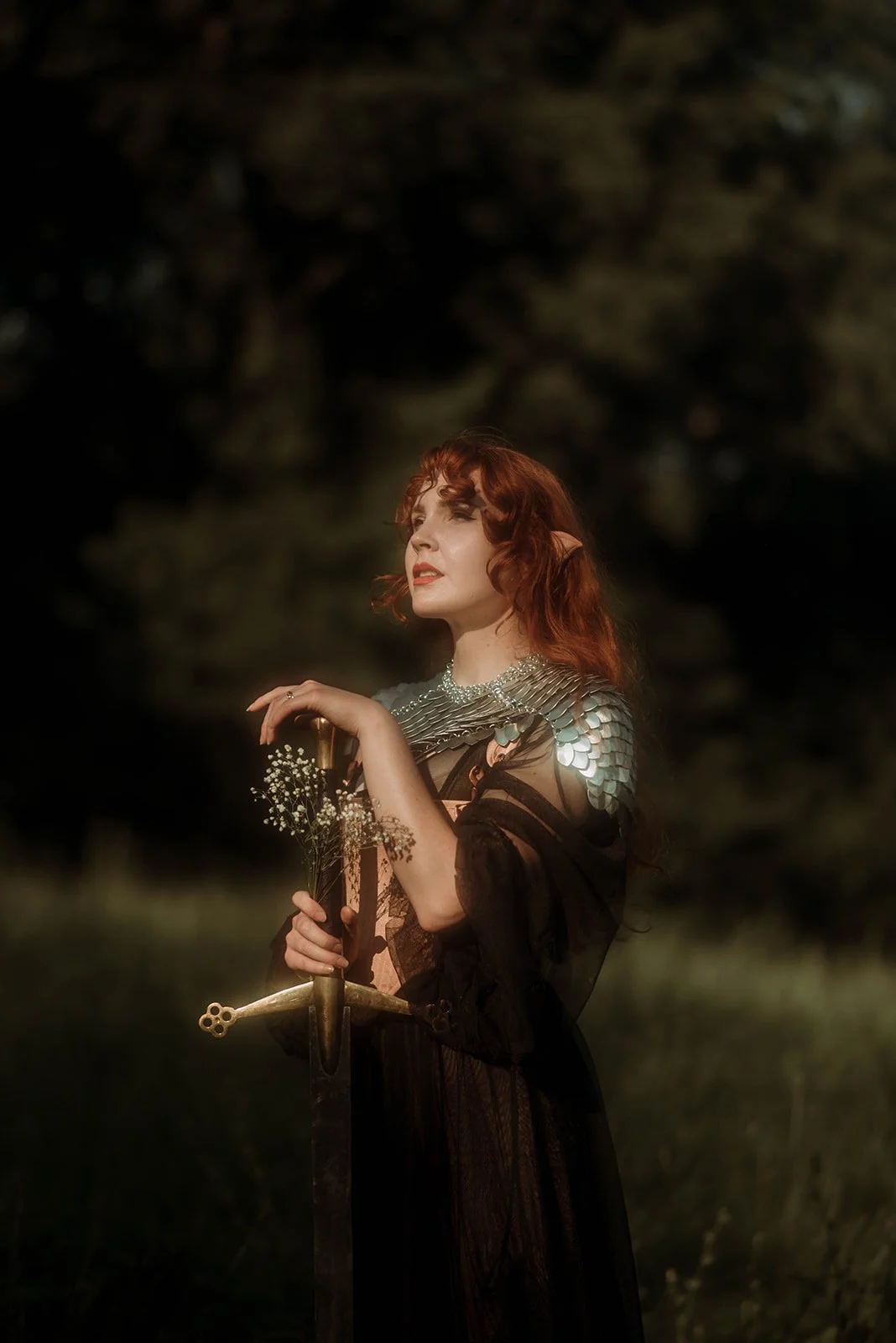 A woman with red hair and pointed ears dressed as an elf holding a sword and flowers outdoors at sunset.