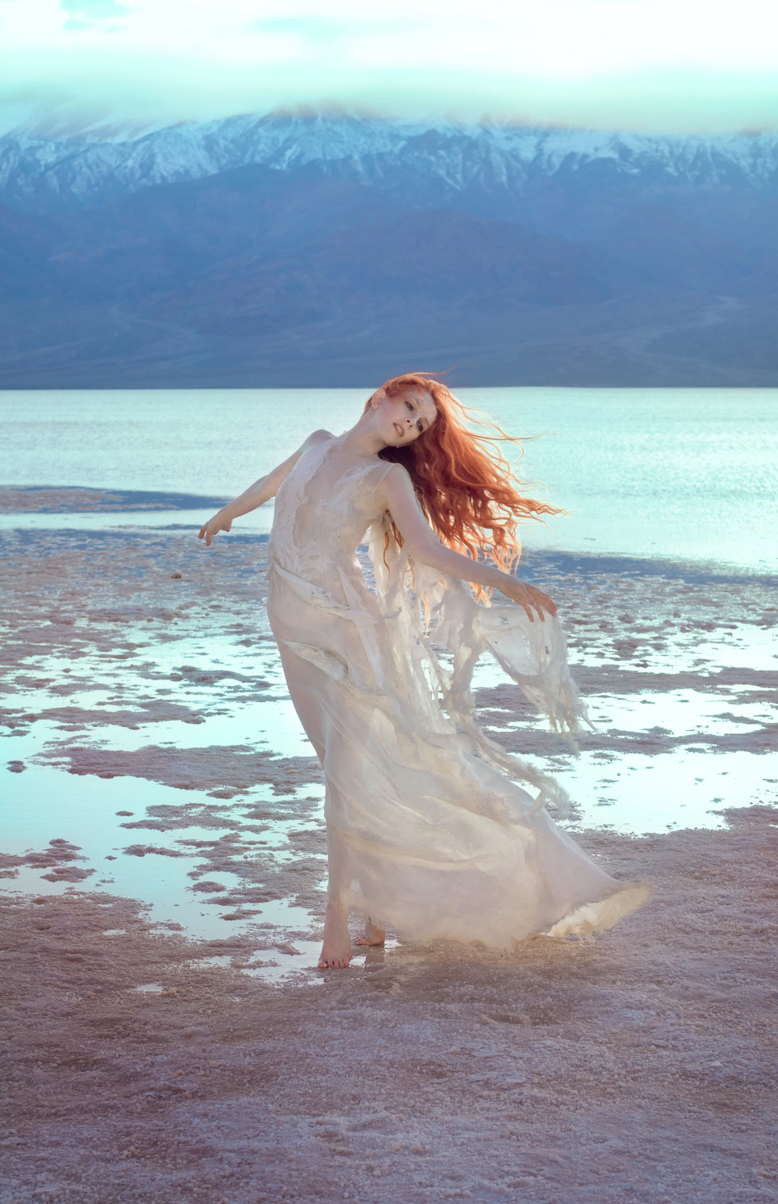 A woman with long red hair dancing barefoot on a wet, textured shoreline near a calm lake, with snow-capped mountains and a cloudy sky in the background.