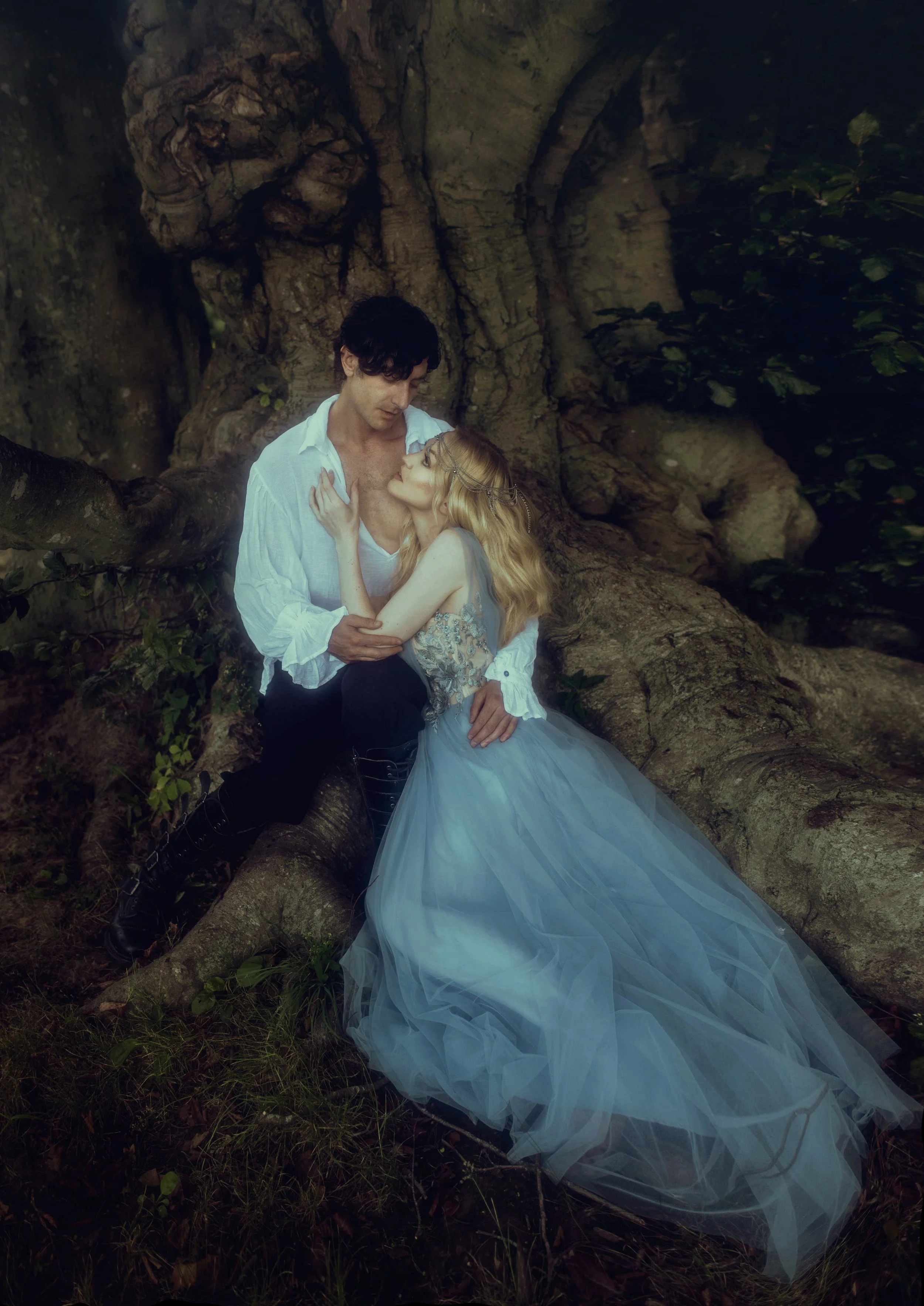 A man and woman dressed in period-style clothing, sitting on a large tree root in a wooded area, gazing into each other's eyes.