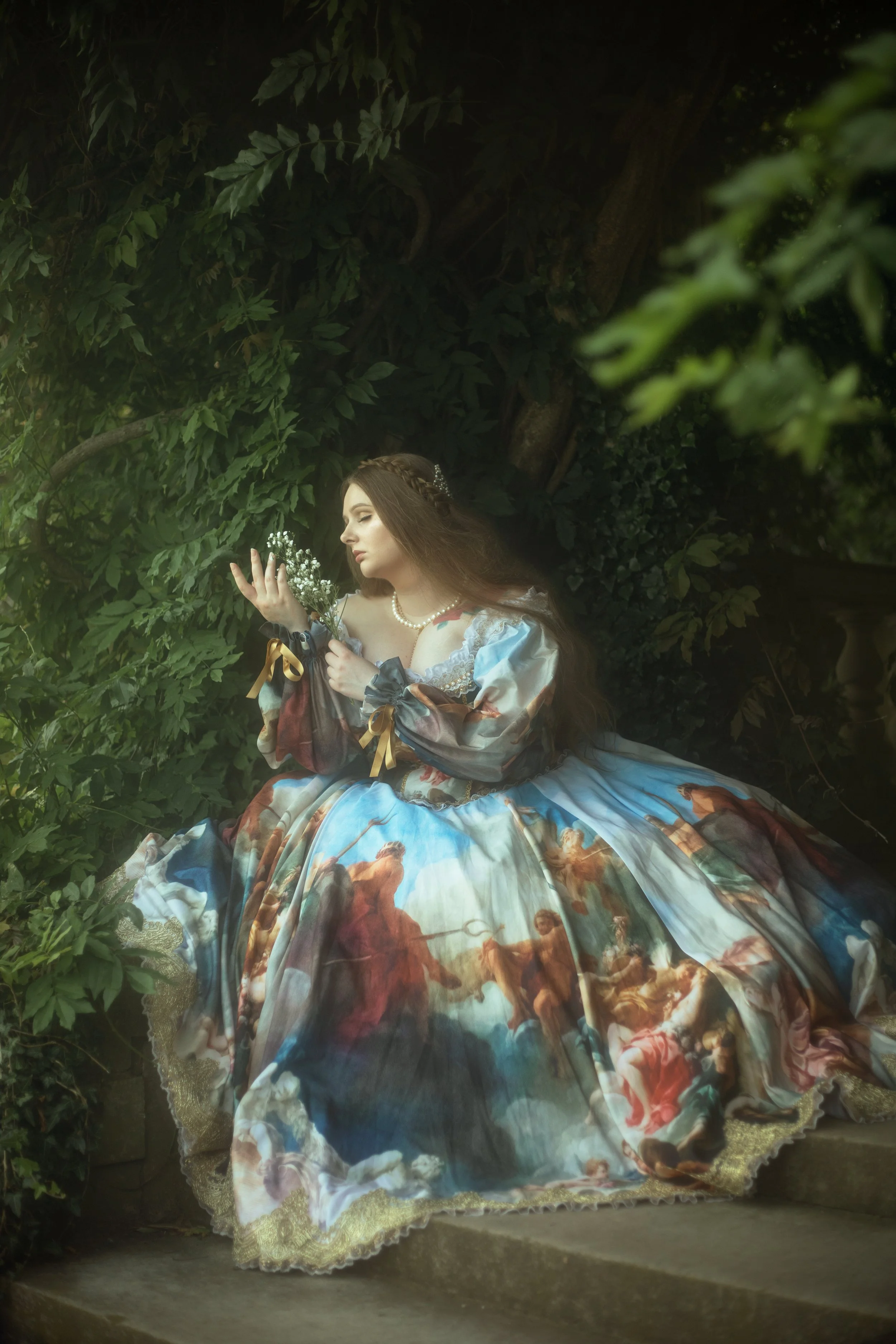 A woman dressed in an elaborate, historical-style gown with a detailed painting pattern, sitting on stone steps near lush green foliage, holding a small bouquet of white flowers.