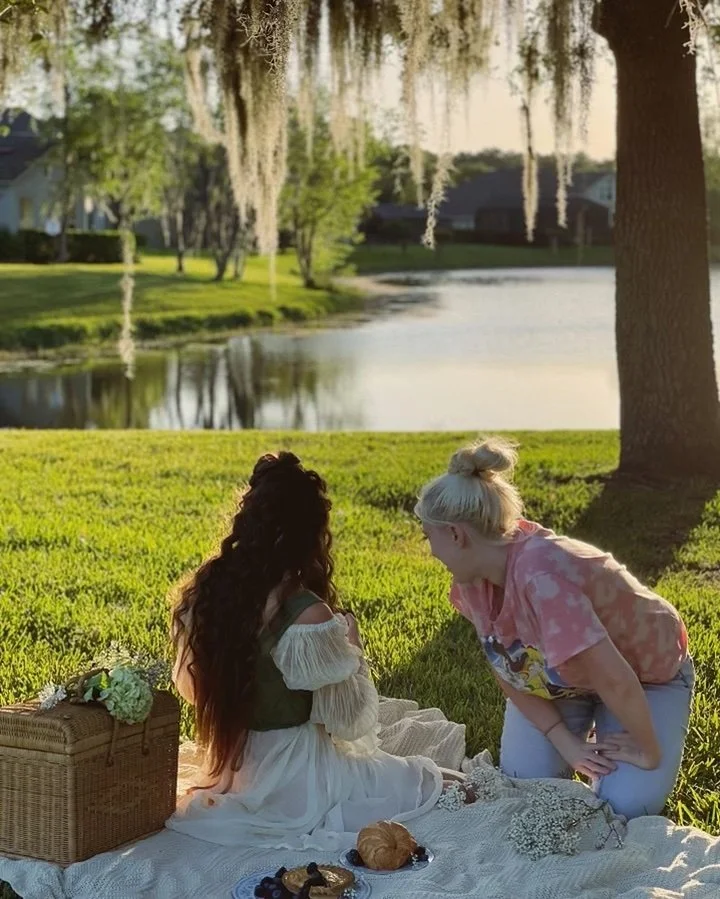 Two women having a picnic by a lake, one with long dark hair sitting on a blanket, the other with blonde hair kneeling, during sunset with trees and water in the background.