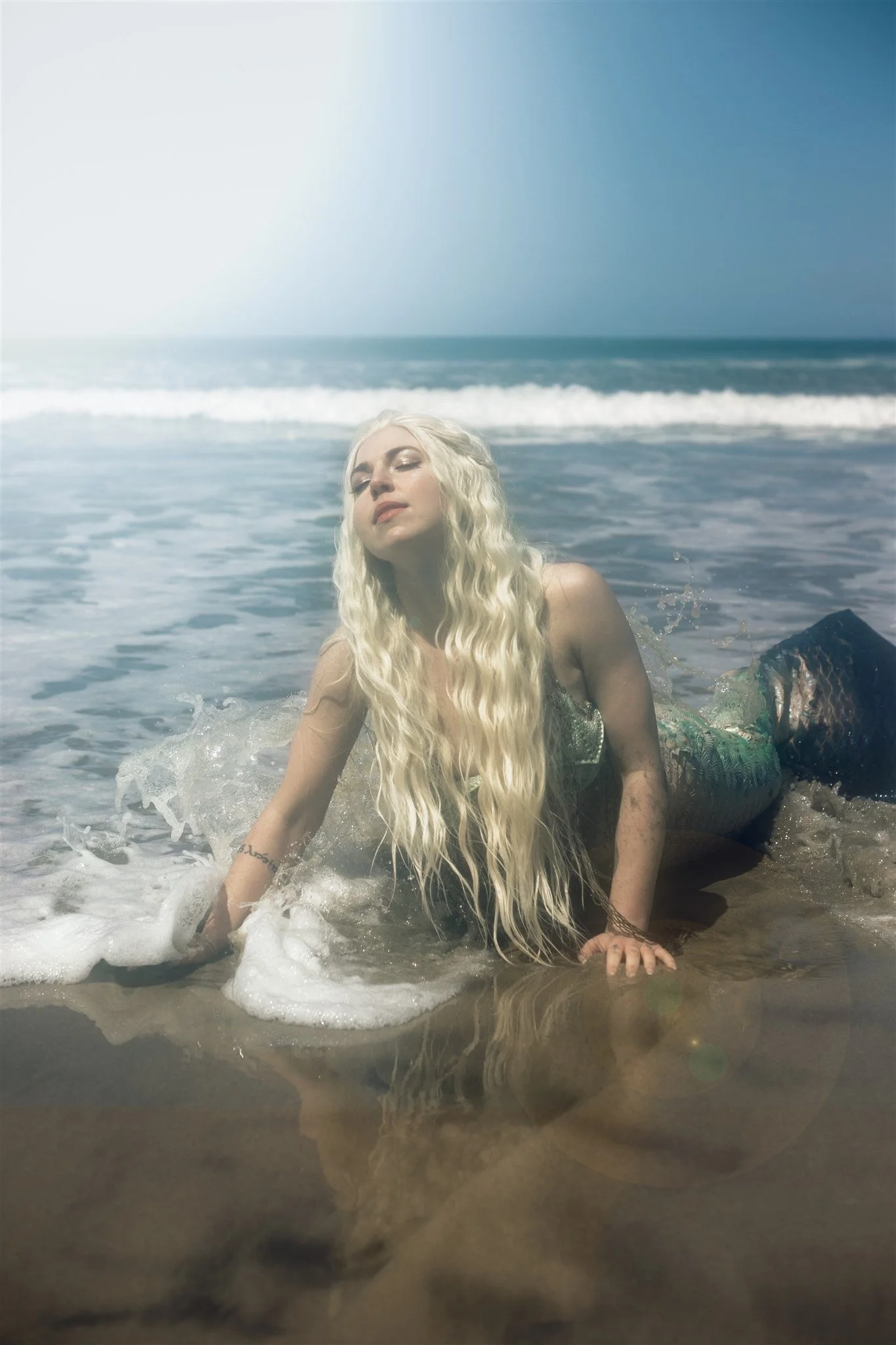 A woman with long blonde hair lying on the beach with her hand on the sand, facing the water with waves in the background and a clear blue sky.