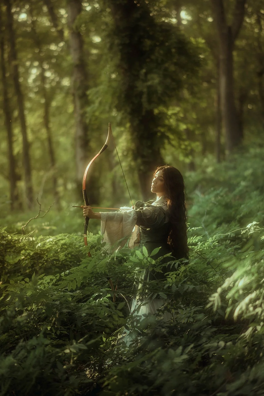 Woman with long dark hair drawing a bow in a green forest during daylight.