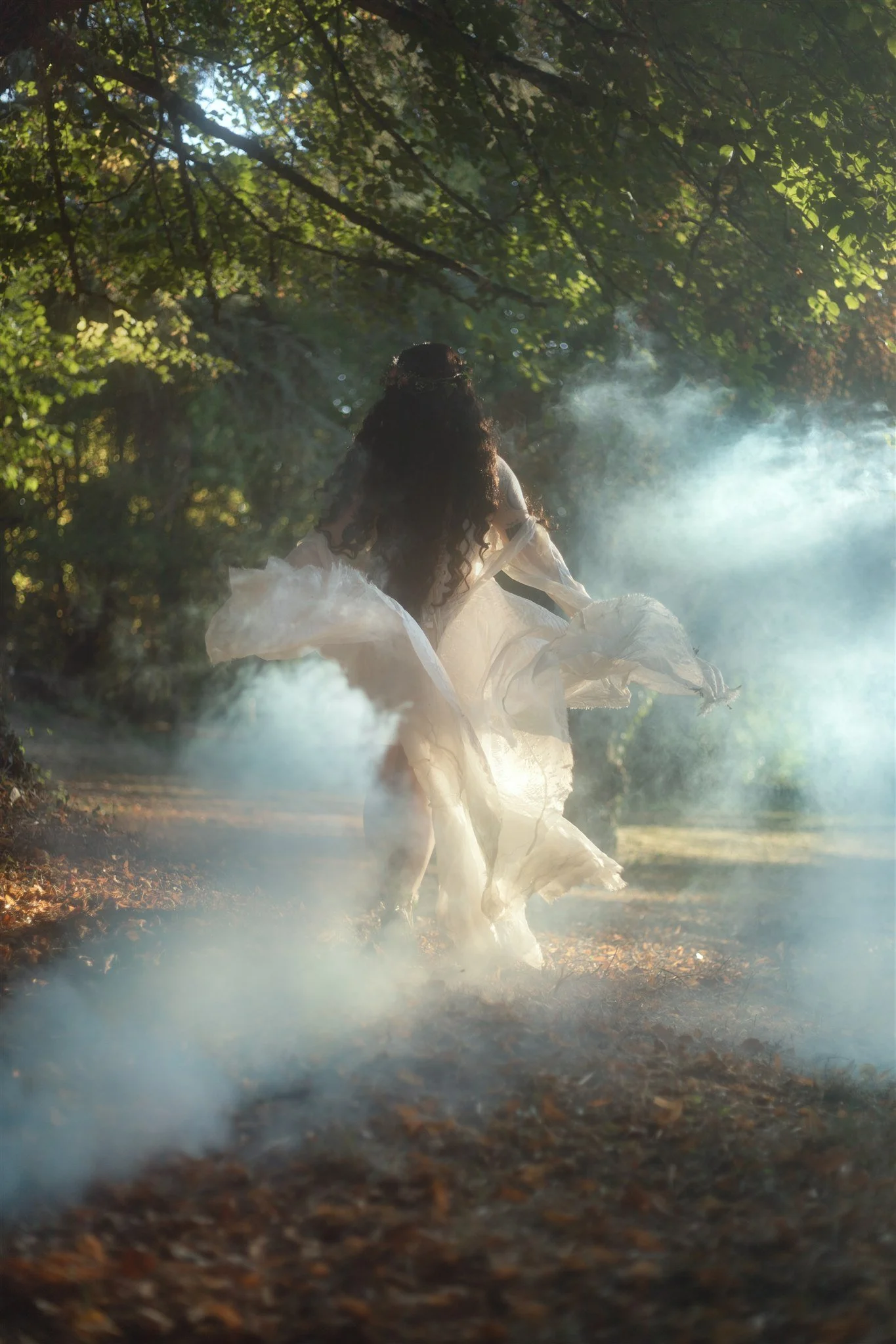A woman in a flowing white dress walking through a foggy forest path surrounded by trees and greenery.