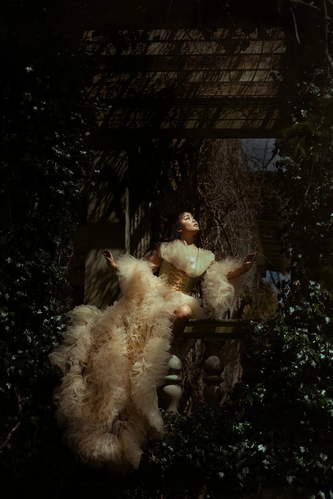 A woman in a flowing, ruffled, beige gown with long, puffed sleeves sits on a balcony surrounded by greenery. She looks upwards with a serene expression, and the scene is illuminated with soft lighting creating a dramatic effect.