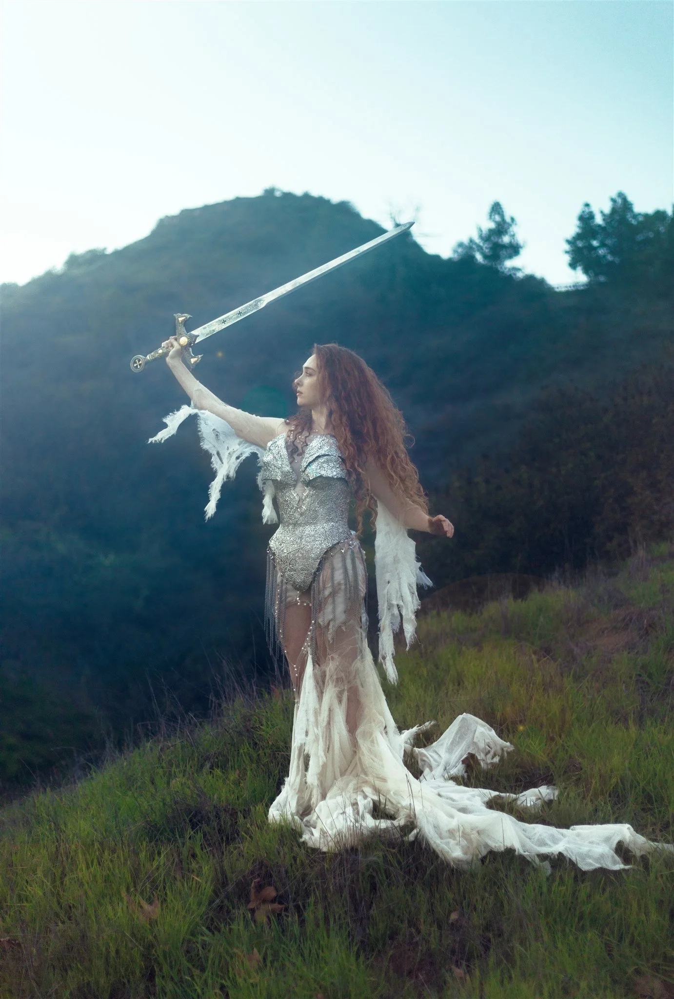 Chappell Roan dressed in elaborate fantasy costume holding a sword above her head in a grassy field with forested hills in the background.