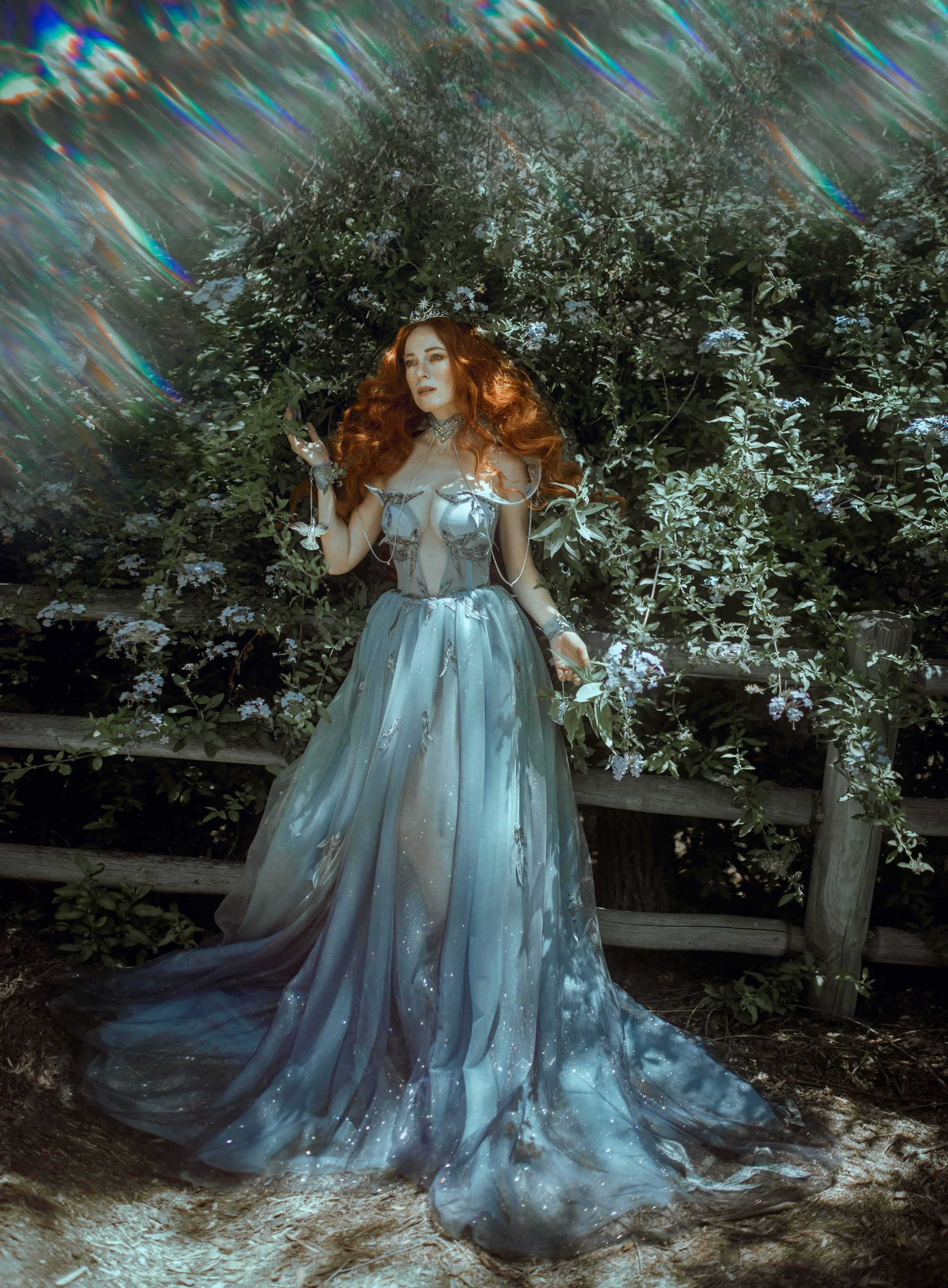 A woman with long red hair wearing a flowing blue fantasy dress standing outdoors in front of green bushes, with a wooden fence behind her.
