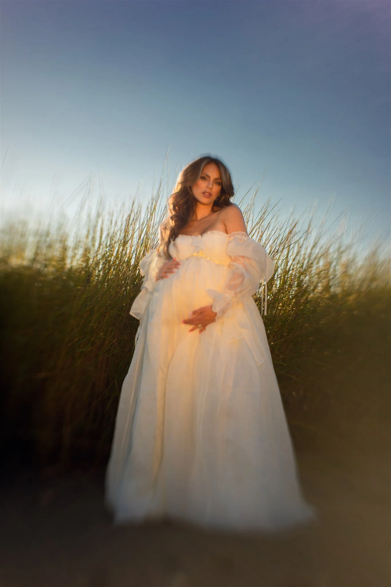 A woman in a flowing, white, off-shoulder dress standing in a field of tall grass with a clear blue sky in the background.