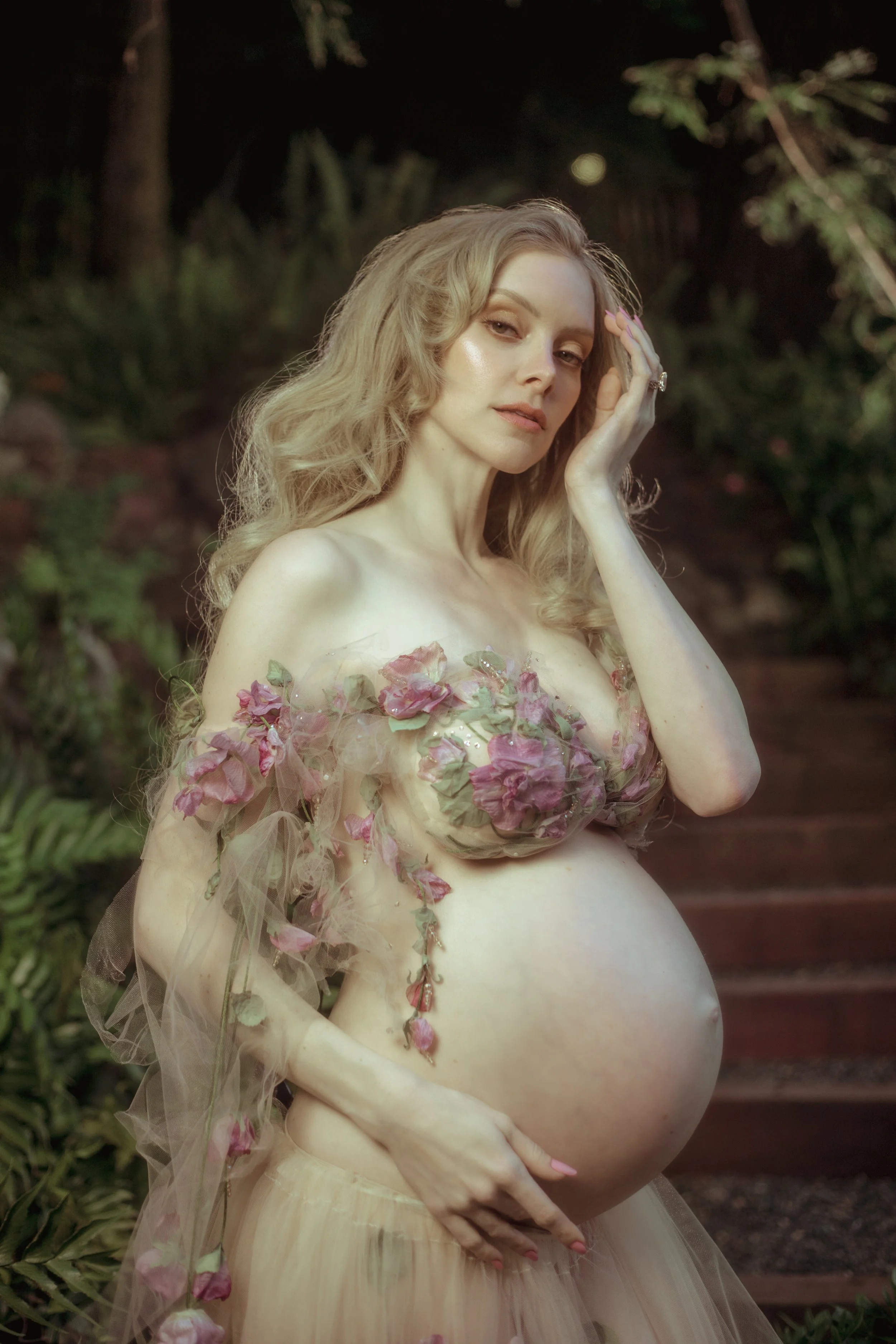 Pregnant woman in a floral-inspired, semi-transparent gown standing outdoors, gently touching her face and stomach, amidst greenery and a wooden staircase.