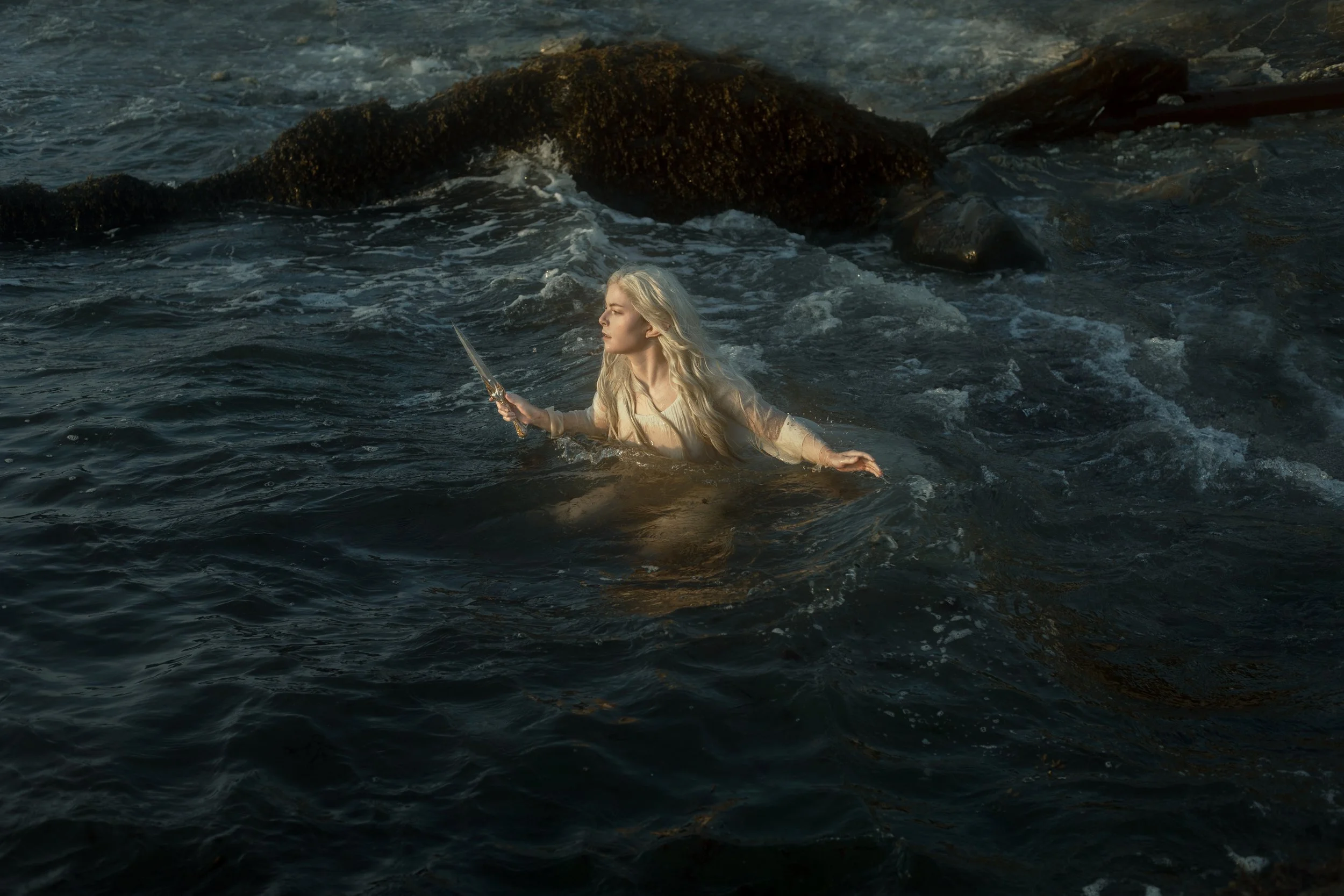 A woman with long blonde hair holding a sword in her right hand, standing in the ocean water near rocks with visible waves and foam.