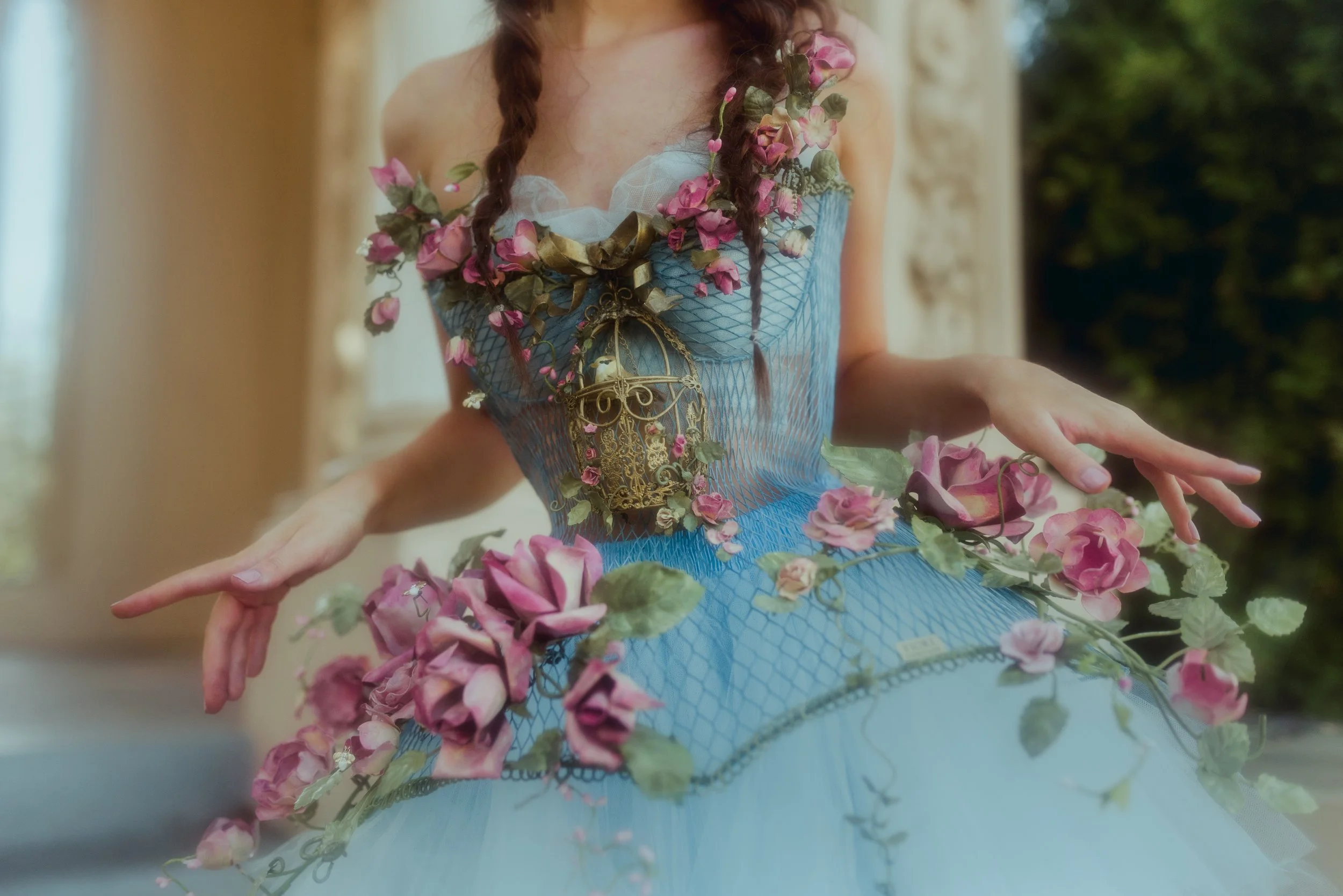 A woman wearing a blue gown decorated with pink flowers and green leaves, with a gold birdcage jewelry piece on her chest, and her hands gracefully extended outward.