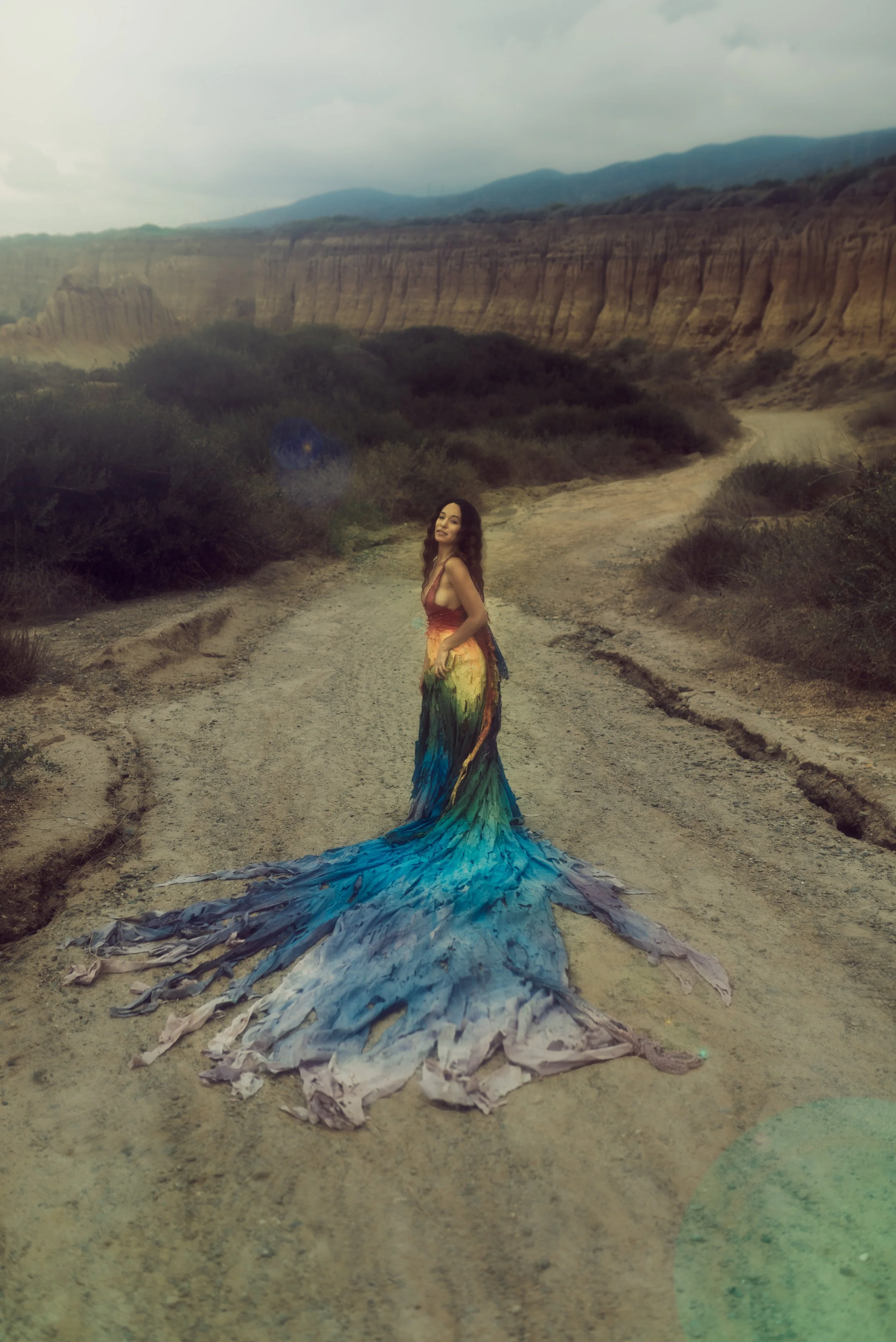 A woman wearing a colorful, rainbow-themed dress with a train stands on a dirt road in a desert landscape with cliffs and mountains in the background.