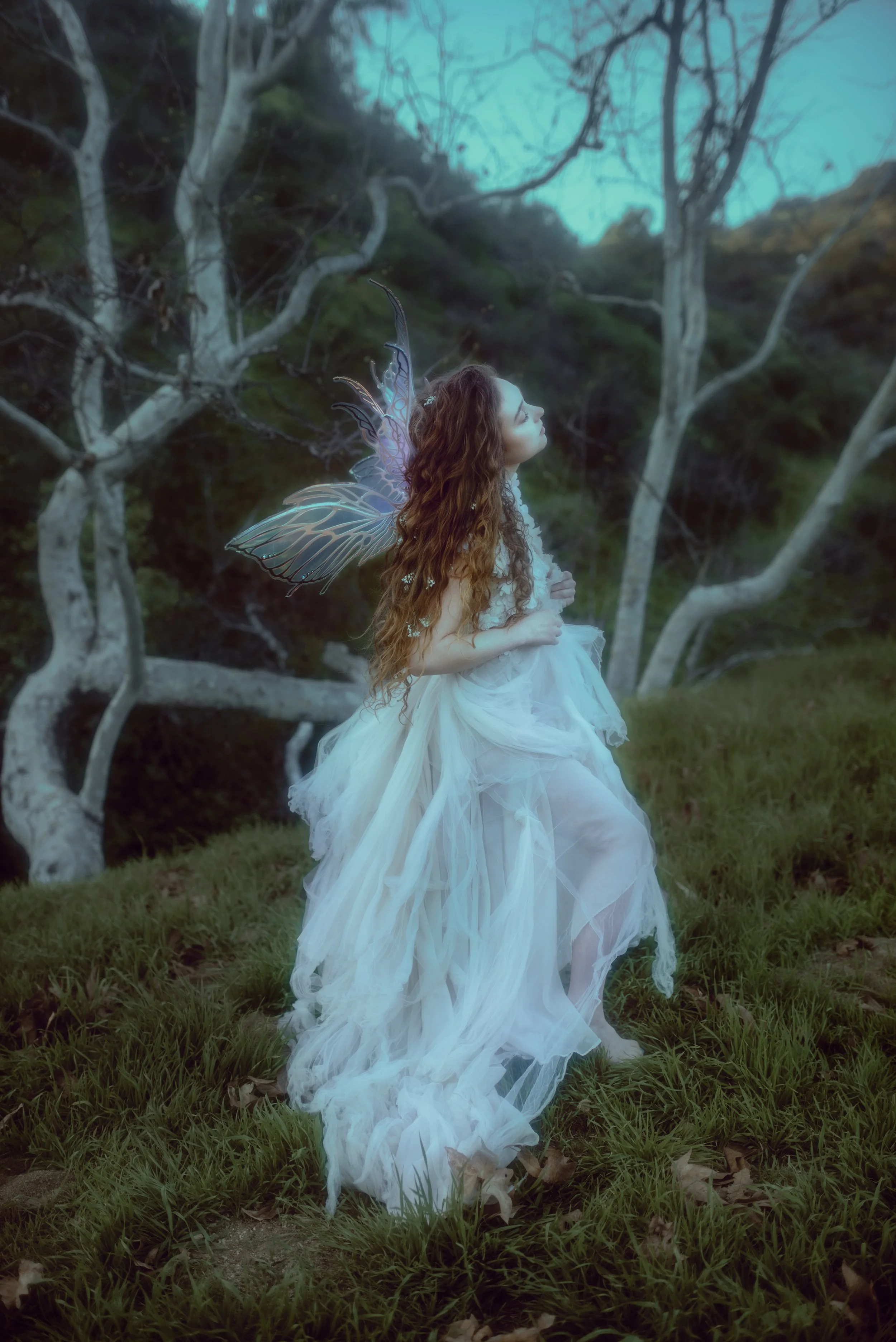 Chappell Roan with fairy wings and long, curly brown hair, dressed in a flowing white gown, standing in a grassy area with leafless trees in the background, looking upwards with a serene expression.