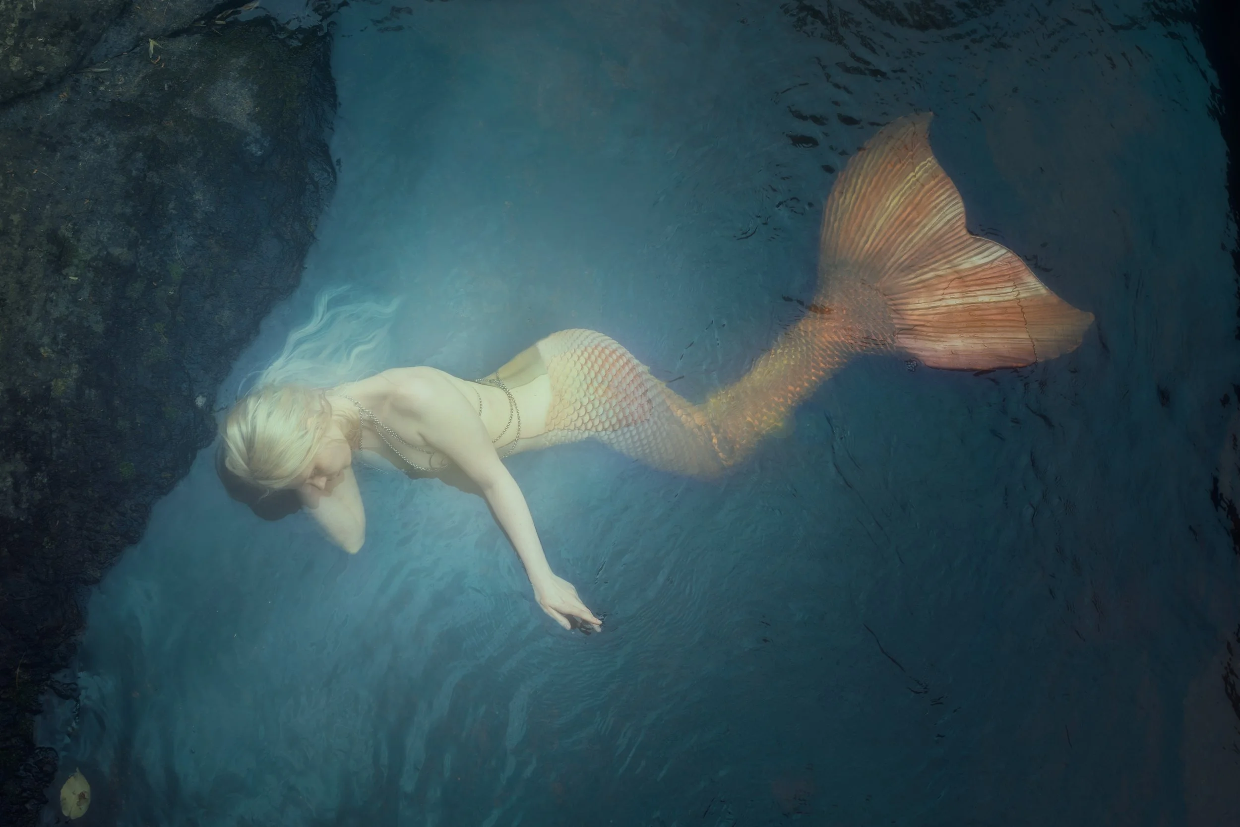 A woman with a mermaid tail floating in a body of water near a rocky area.