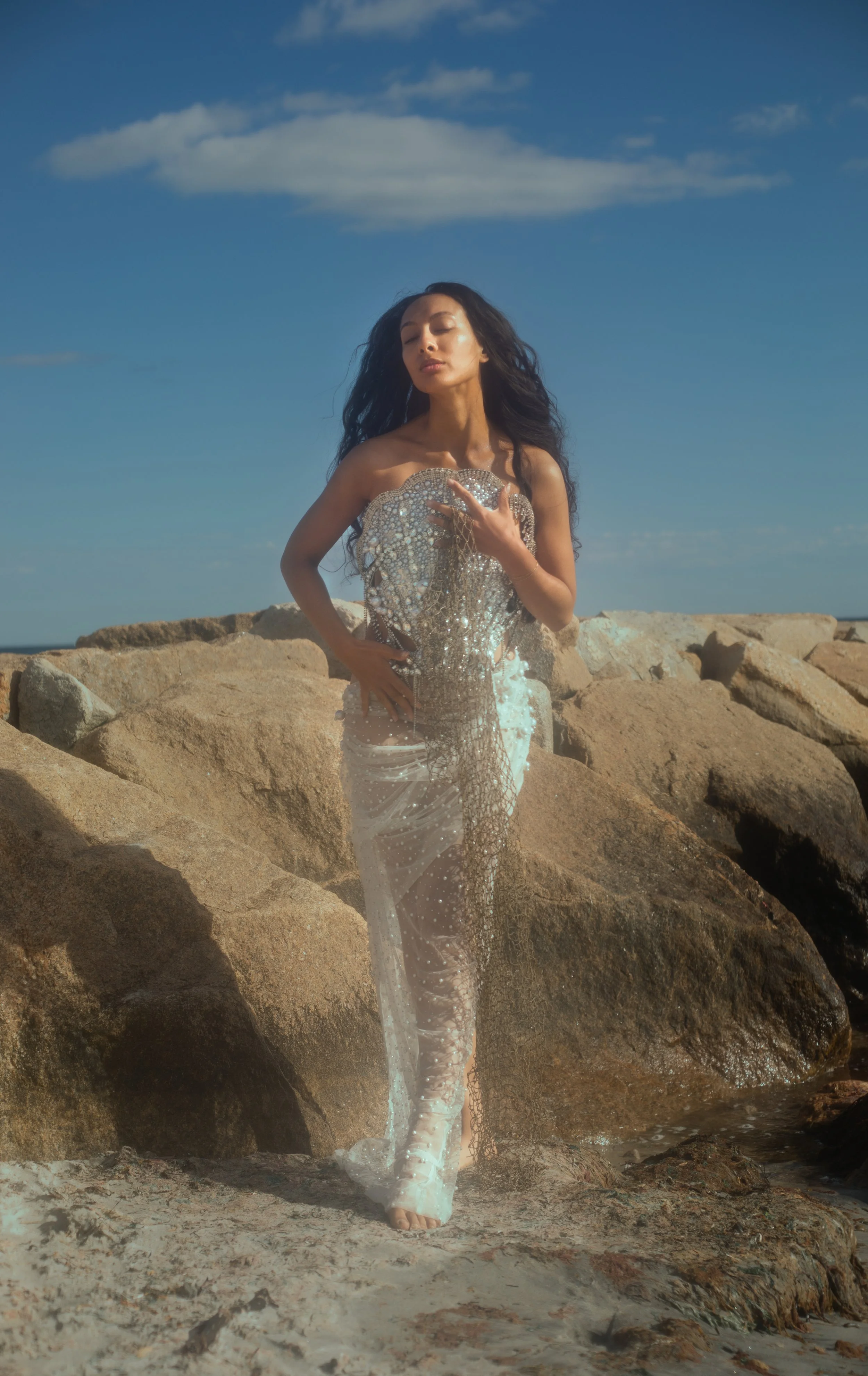 A woman standing on rocks at the beach wearing a shimmering, elegant dress.