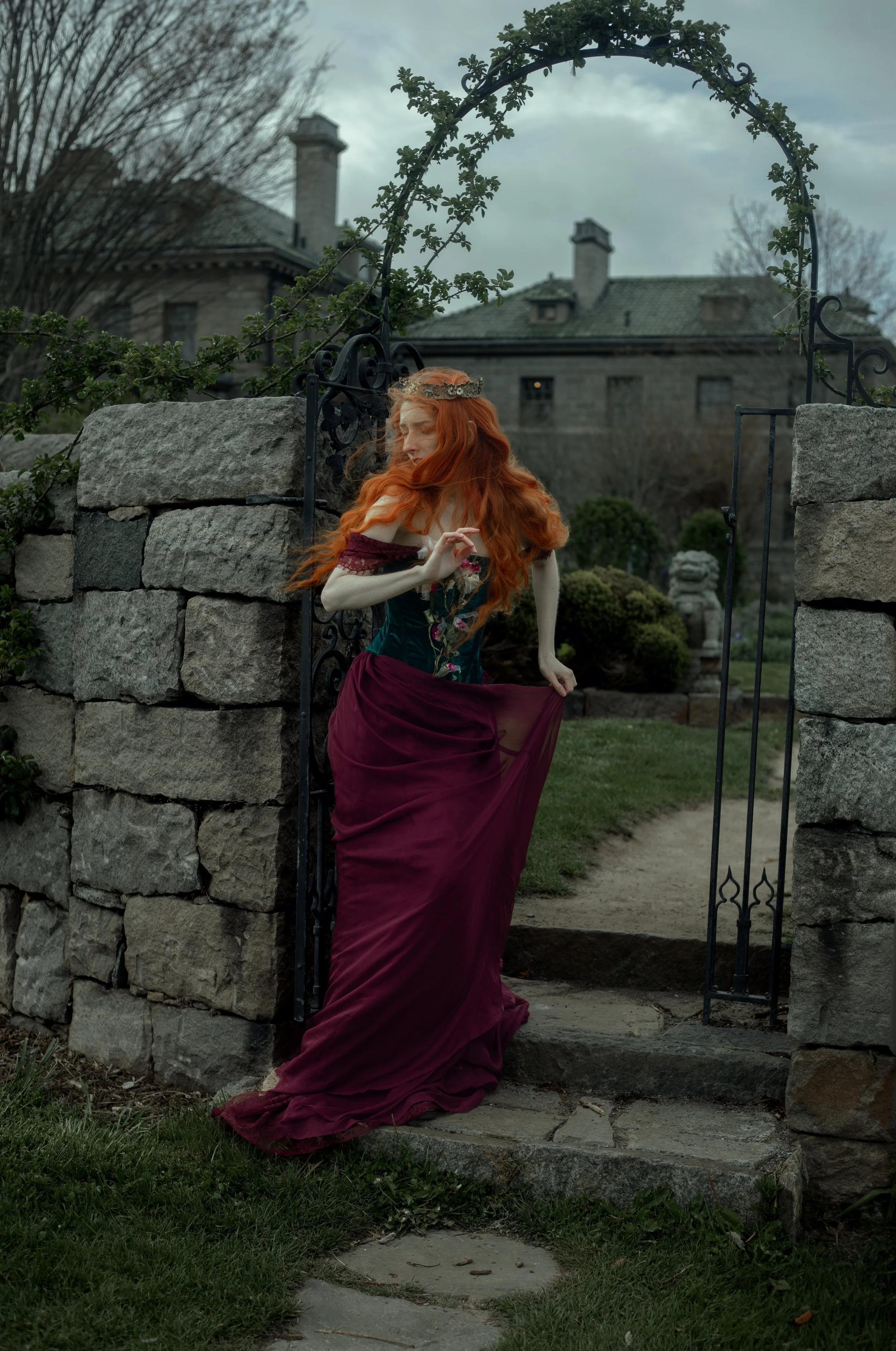 A woman with long red hair in a medieval-style dress standing at an open stone gate on a garden path, with a large castle-like house in the background, overcast skies.