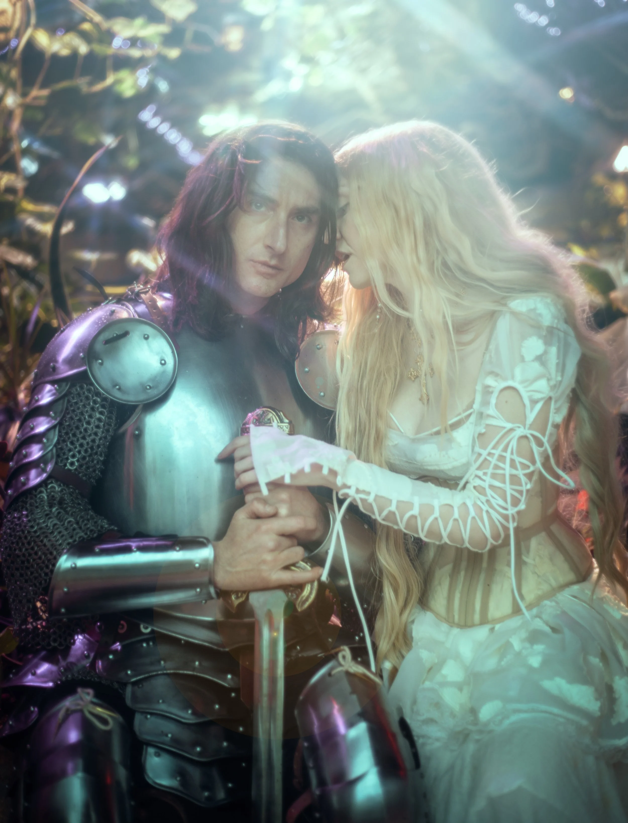A man dressed as a knight in armor with long dark hair and a woman with long blonde hair in a white dress whispering next to him in a forest.